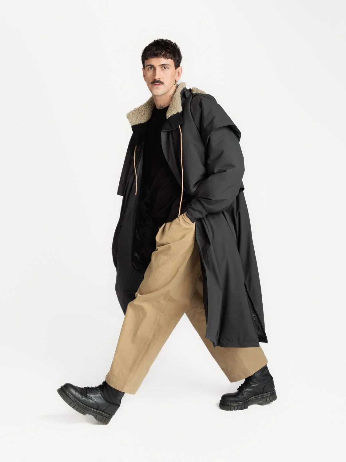 En mann ikledd en lang, svart kraftig kåpe med pelsfór, beige bukser og sorte støvler går i en lett pose som ser ut til å være med i en fottøy- eller moteshoot.
