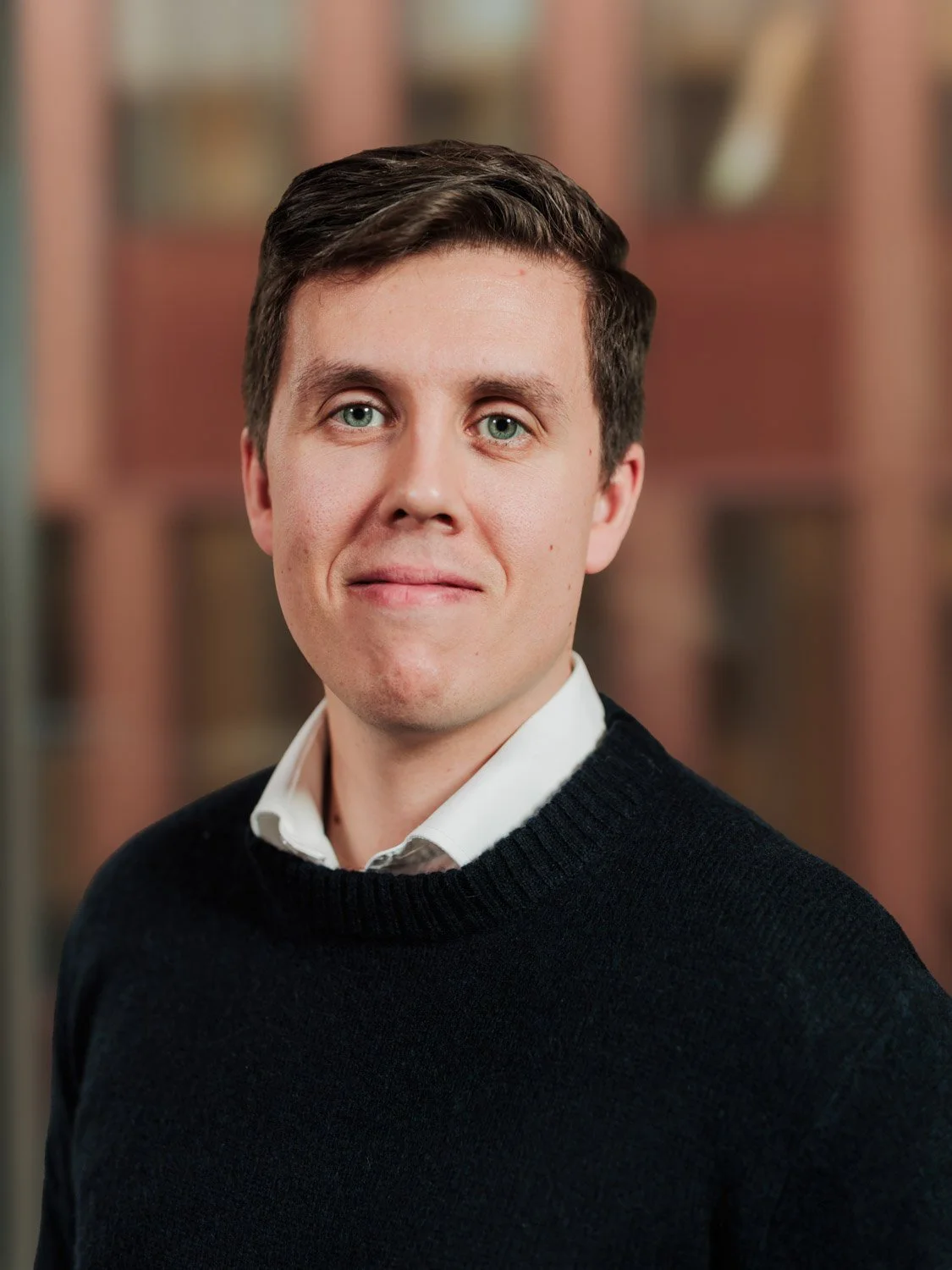 Portrett av en ung mann med mørkt hår, hvit skjorte og svart genser, med bokhylle i bakgrunnen.