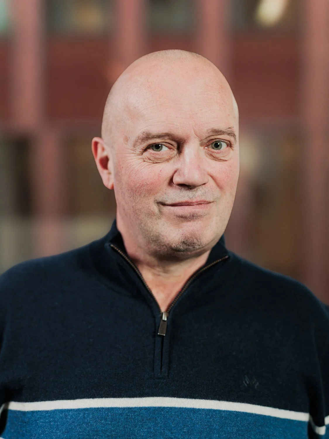 Portrettbilde av en mann med blankskallet hode, iført en mørk genser med en hvit og blå stripe, mot en uskarp, bokhylles-aktig bakgrunn.
