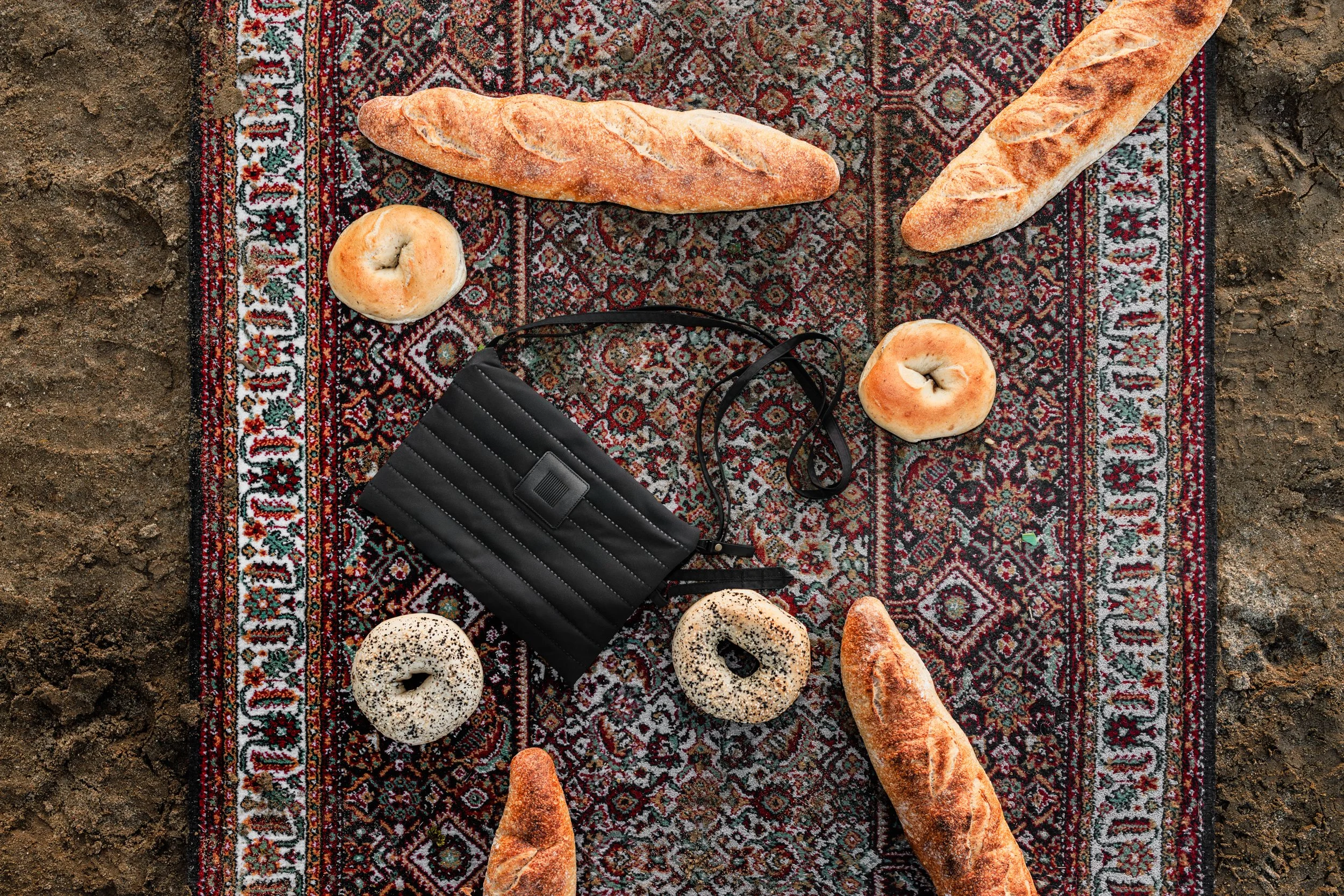 Brød og bagels plassert på et teppe på bakken med jord rundt, samt en svart veske.