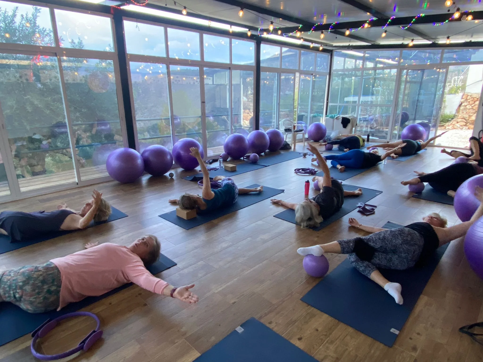 People participating in a yoga class in a sunlit room with large windows, purple exercise balls, and string lights overhead, some using yoga blocks and mats.