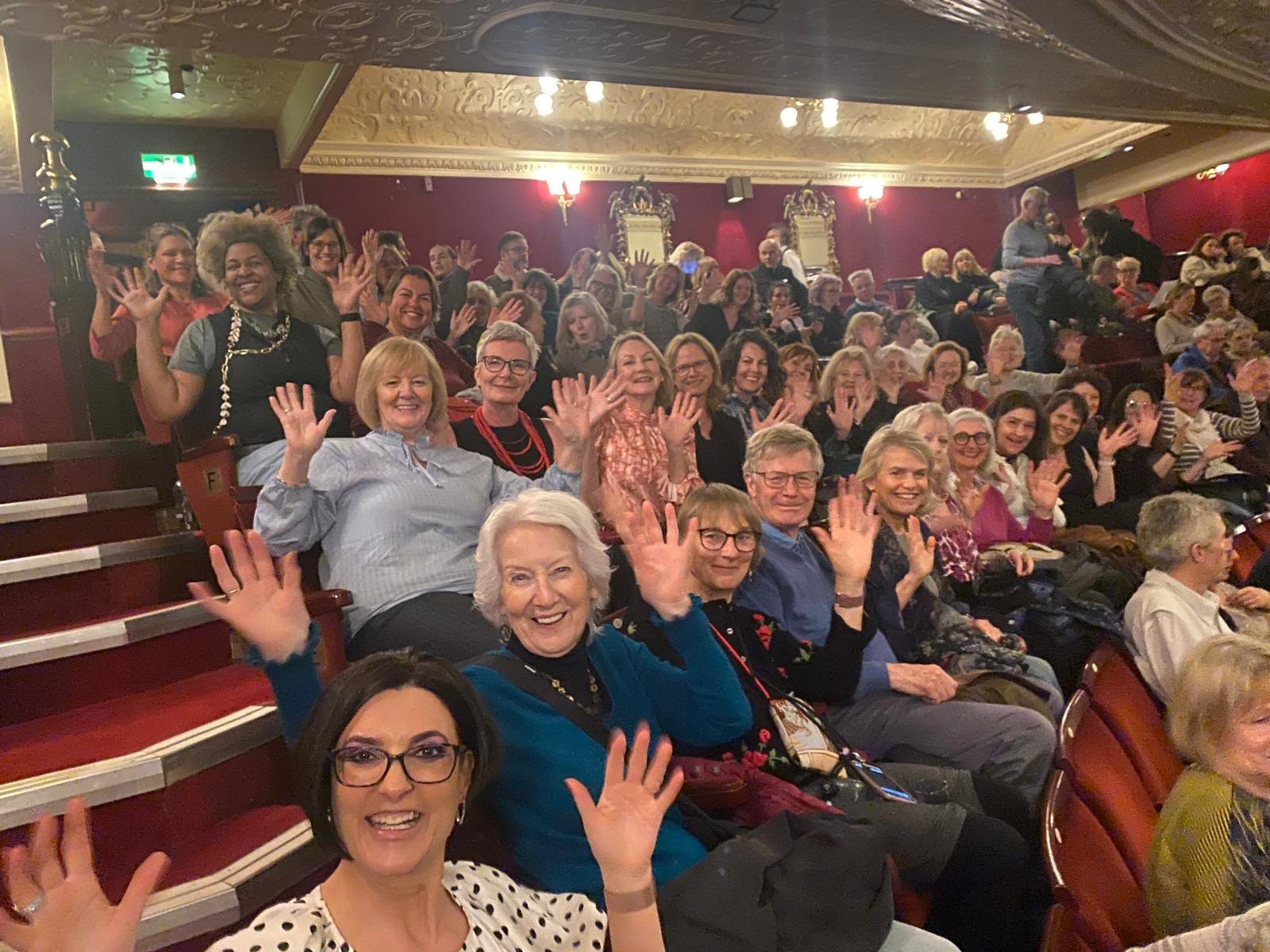 Group of people sitting in theater seats, smiling, waving, and looking at the camera, with ornate ceiling and walls in the background.