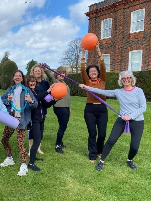 Group of women outdoors holding orange Pilates balls and equipment, standing on grass in front of a large brick building under partly cloudy sky.