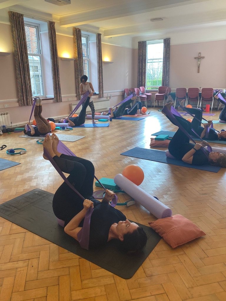 A group of people participating in a yoga or Pilates class, lying on yoga mats, with resistance bands, pillows, and exercise balls, practicing exercises in a bright room with large windows and curtains.