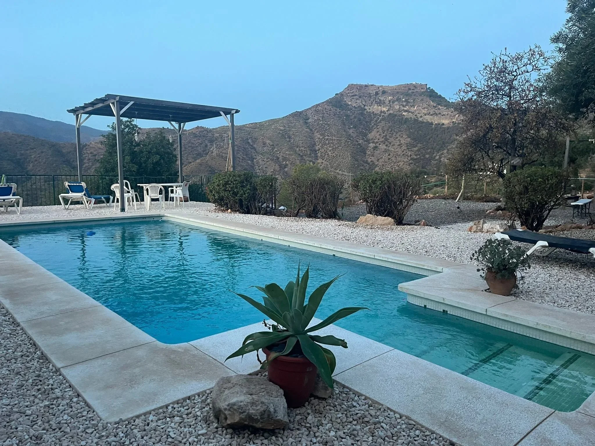 An outdoor swimming pool surrounded by gravel and potted plants, with a mountain view in the background. There are lounge chairs and a shaded seating area with chairs and a table.
