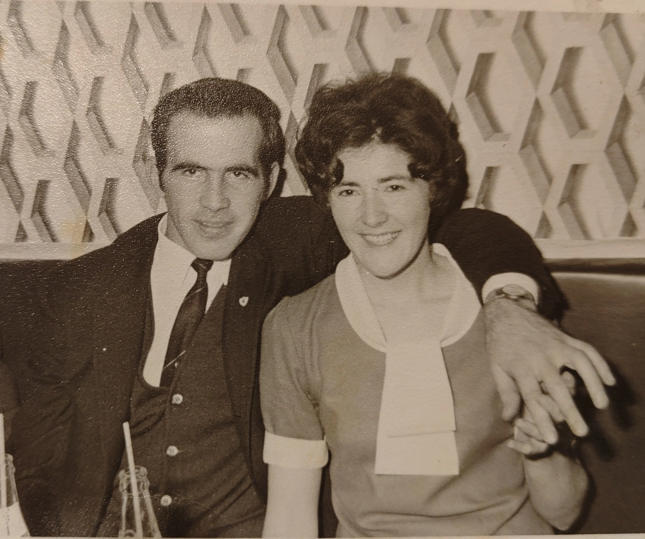 A black and white photo of a man and a woman sitting at a table, both smiling at the camera. The man has dark hair and is wearing a suit and tie, while the woman has curled hair and is dressed in a patterned blouse with a tie. The man has his arm around the woman's shoulder. There are bottles and drinking straws on the table, and a decorative lattice wall in the background.