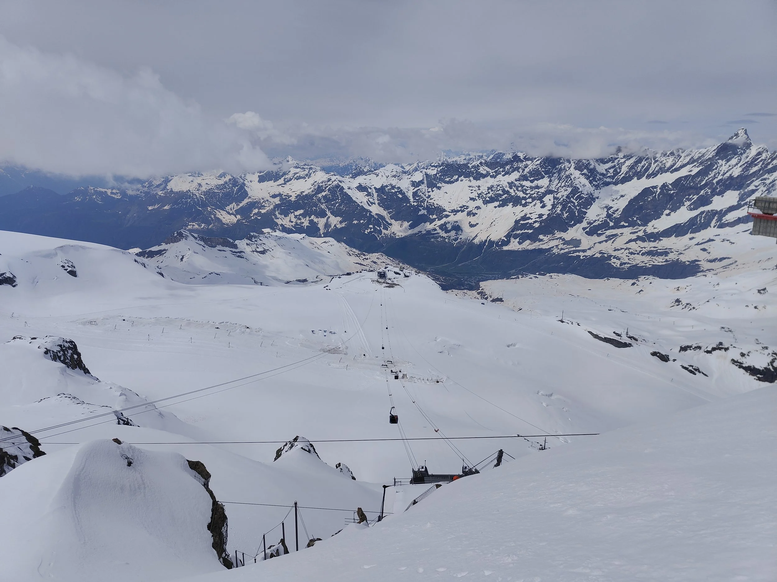 Snow-covered mountain landscape with ski lift cables and towers in the foreground, and distant snow-capped peaks under cloudy skies.