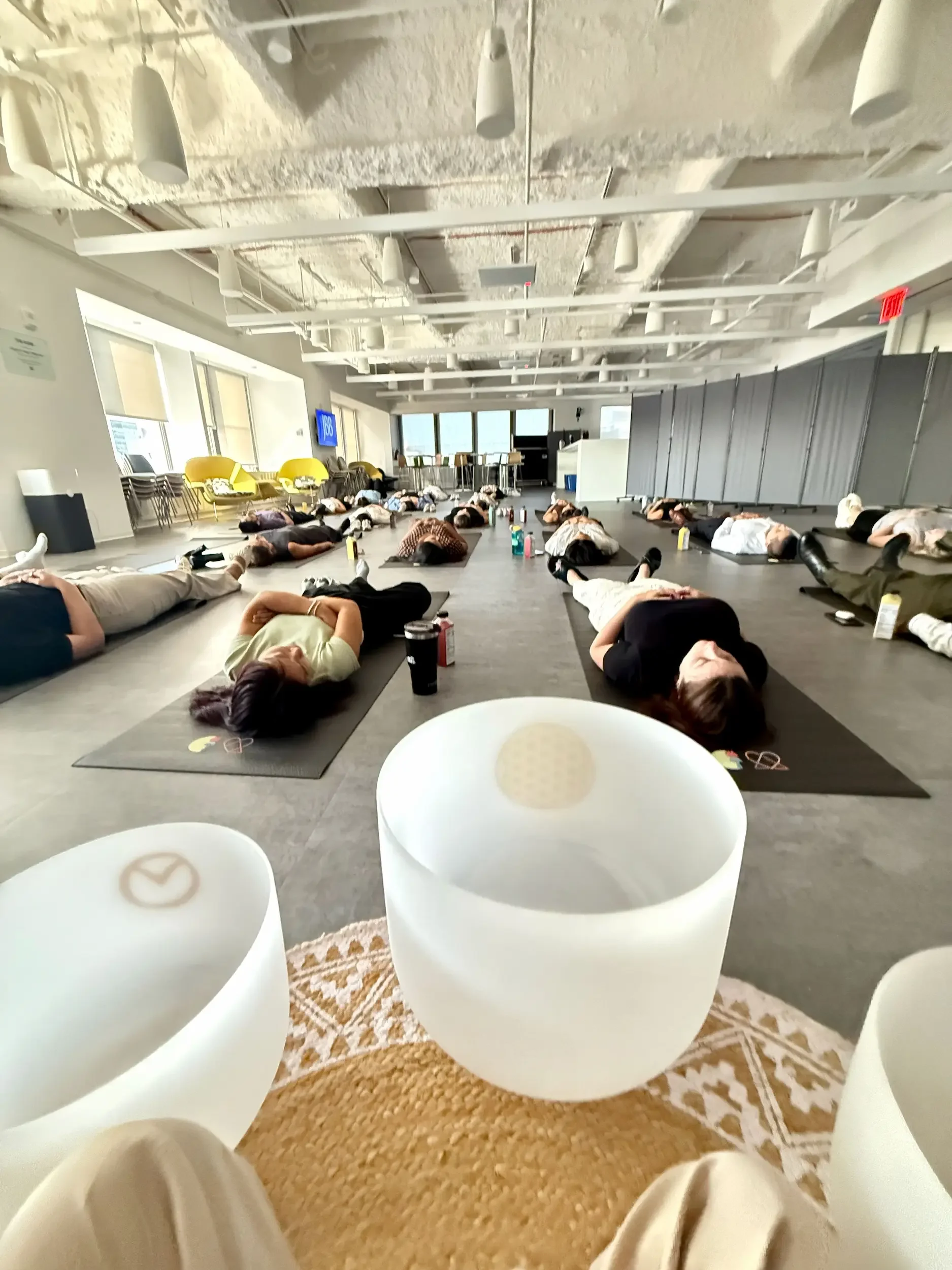 Natalie Chan leading a corporate sound bath for JB&B employees in a modern office space, with participants resting on black yoga mats