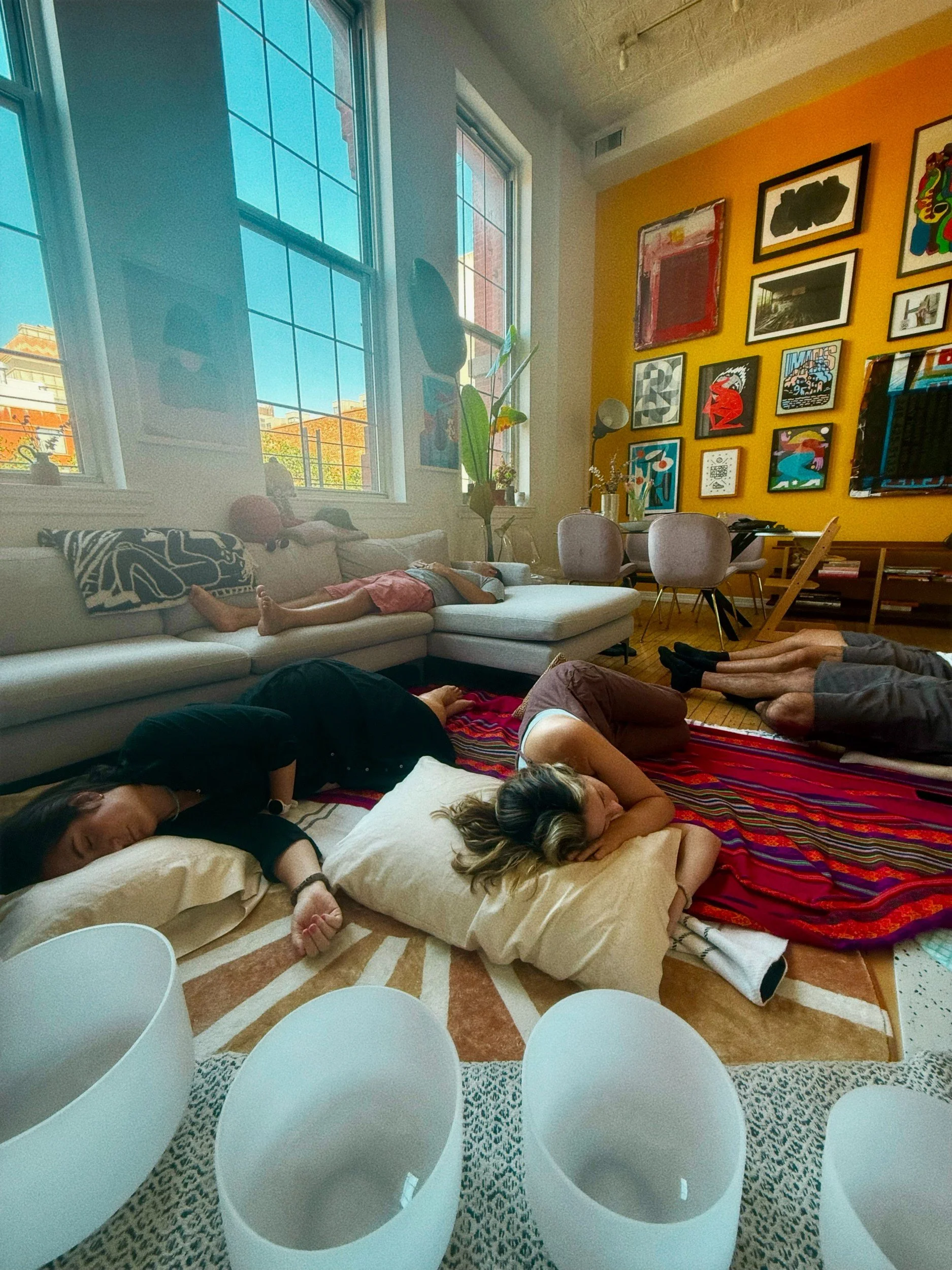 A private at-home sound bath by Soundawn — guests resting on cushions in a sunlit Brooklyn apartment, with crystal singing bowls in the foreground.
