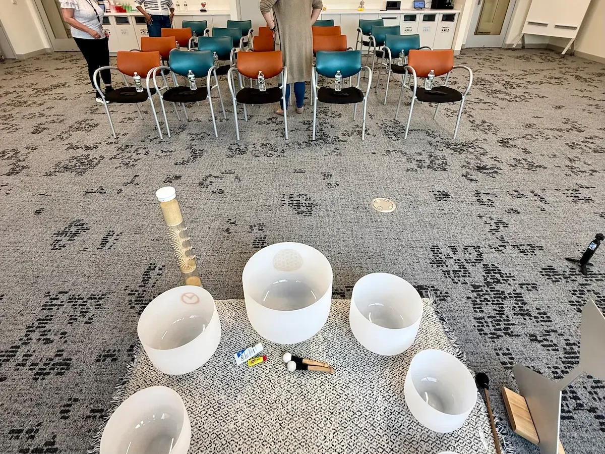 Crystal singing bowls, ocean drum, and chime set up on a woven mat before a corporate sound bath for the Gilead Sciences team at their New Jersey office, with chairs arranged for employees.