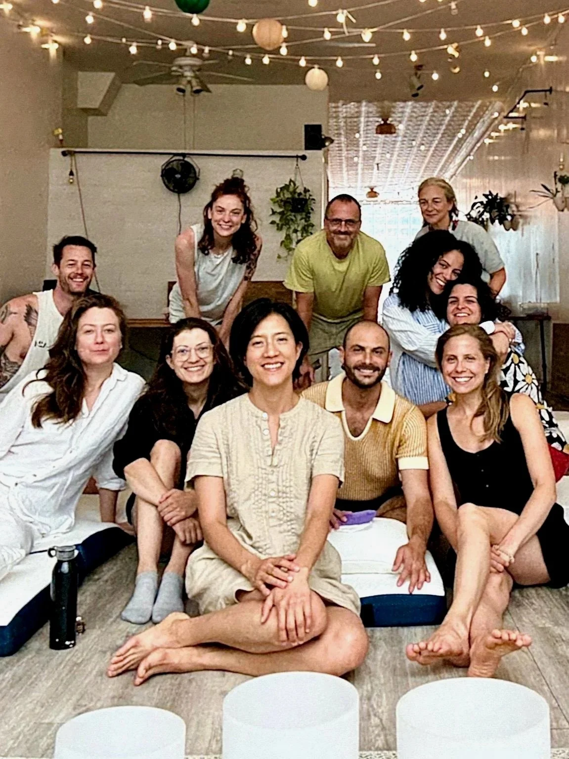 Group of people smiling and sitting together on floor cushions during a community sound bath session, with warm string lights creating a cozy atmosphere.          Ask ChatGPT
