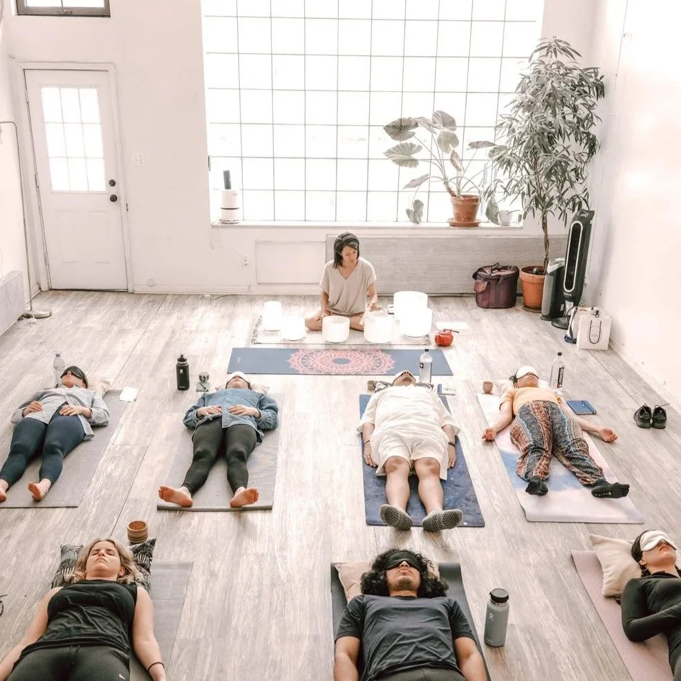 Group lying on yoga mats in deep rest while Natalie Chan plays crystal singing bowls during a sound bath meditation at the sunlit Soundawn studio in Brooklyn.