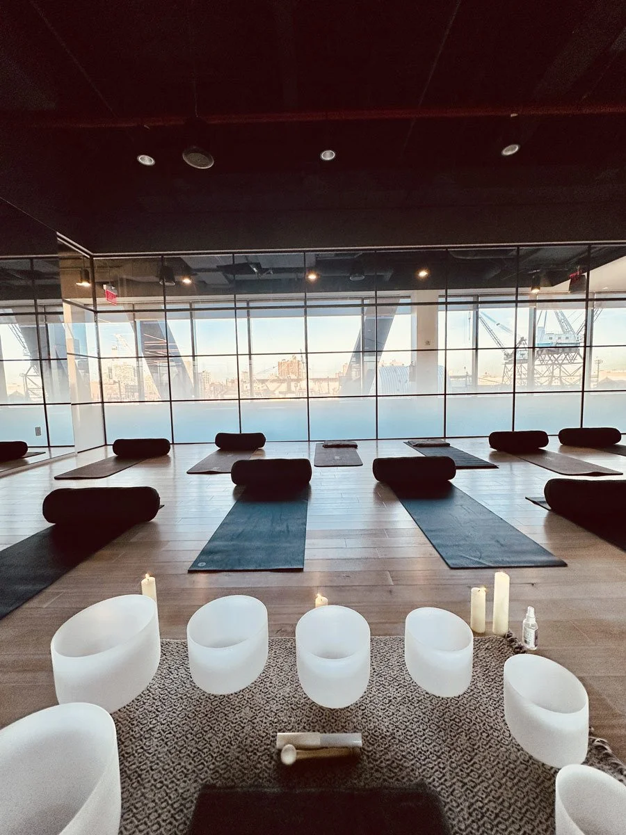 Yoga studio with black mats and bolsters on a wooden floor, large windows with a cityscape view, and white crystal singing bowls with candles and a mallet in the foreground.