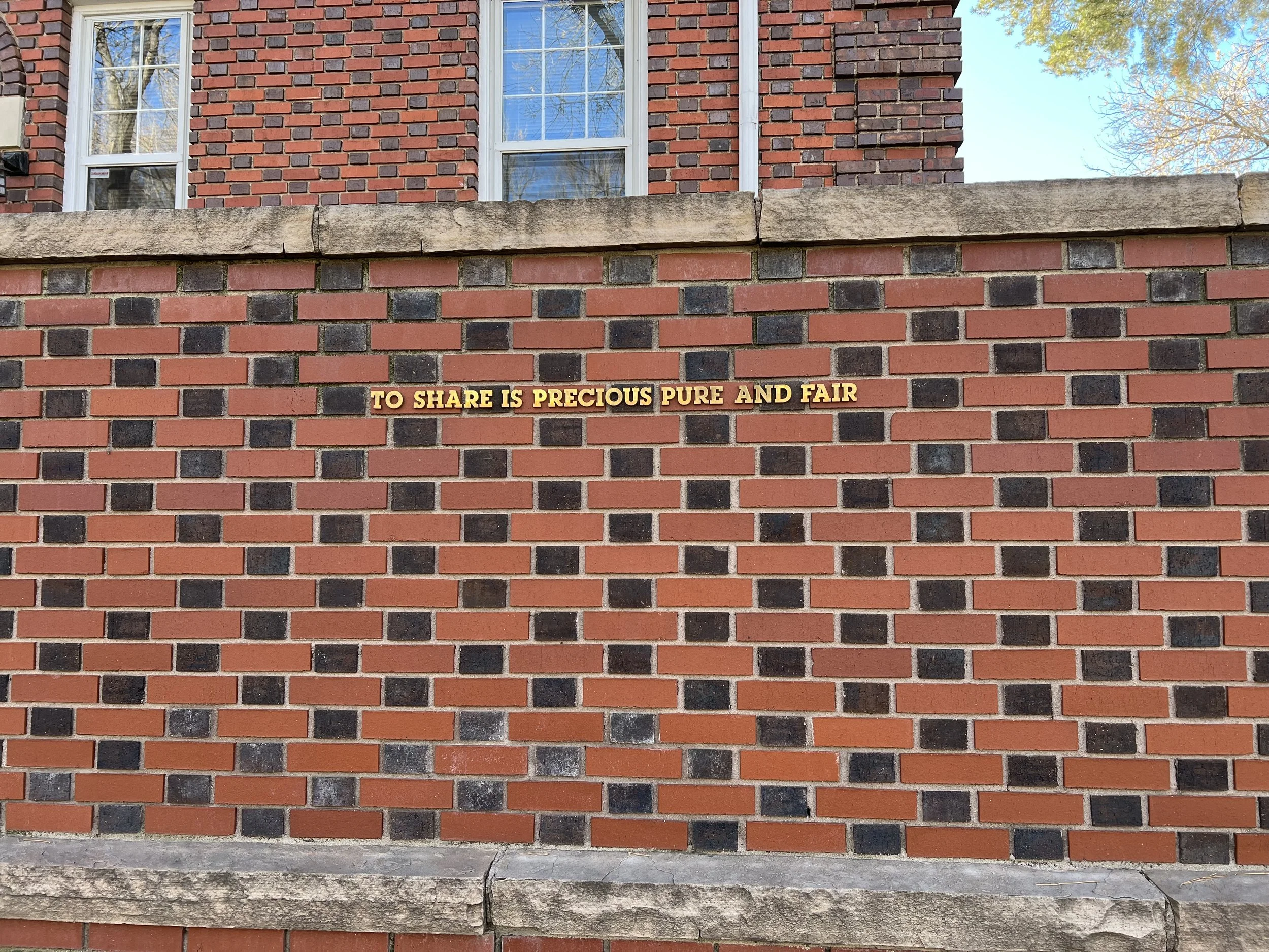 Brick wall with text in gold that says "TO SHARE IS PRECIOUS PURE AND FAIR".