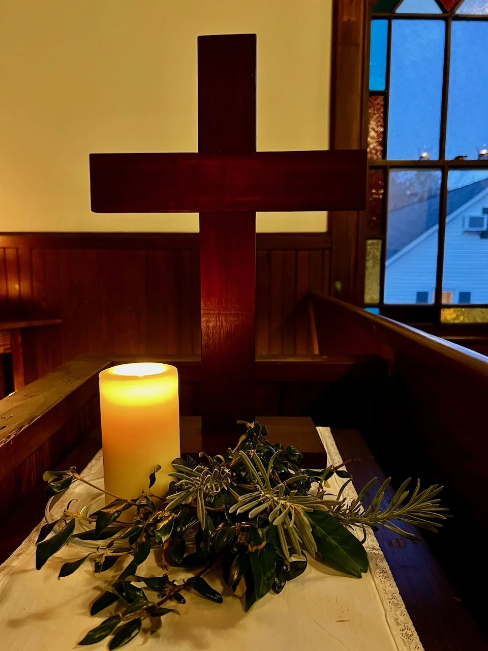 A wooden church cross decorated with greenery, a large cream-colored candle, and a white cloth on a table inside a church, near a window showing a blue evening sky.
