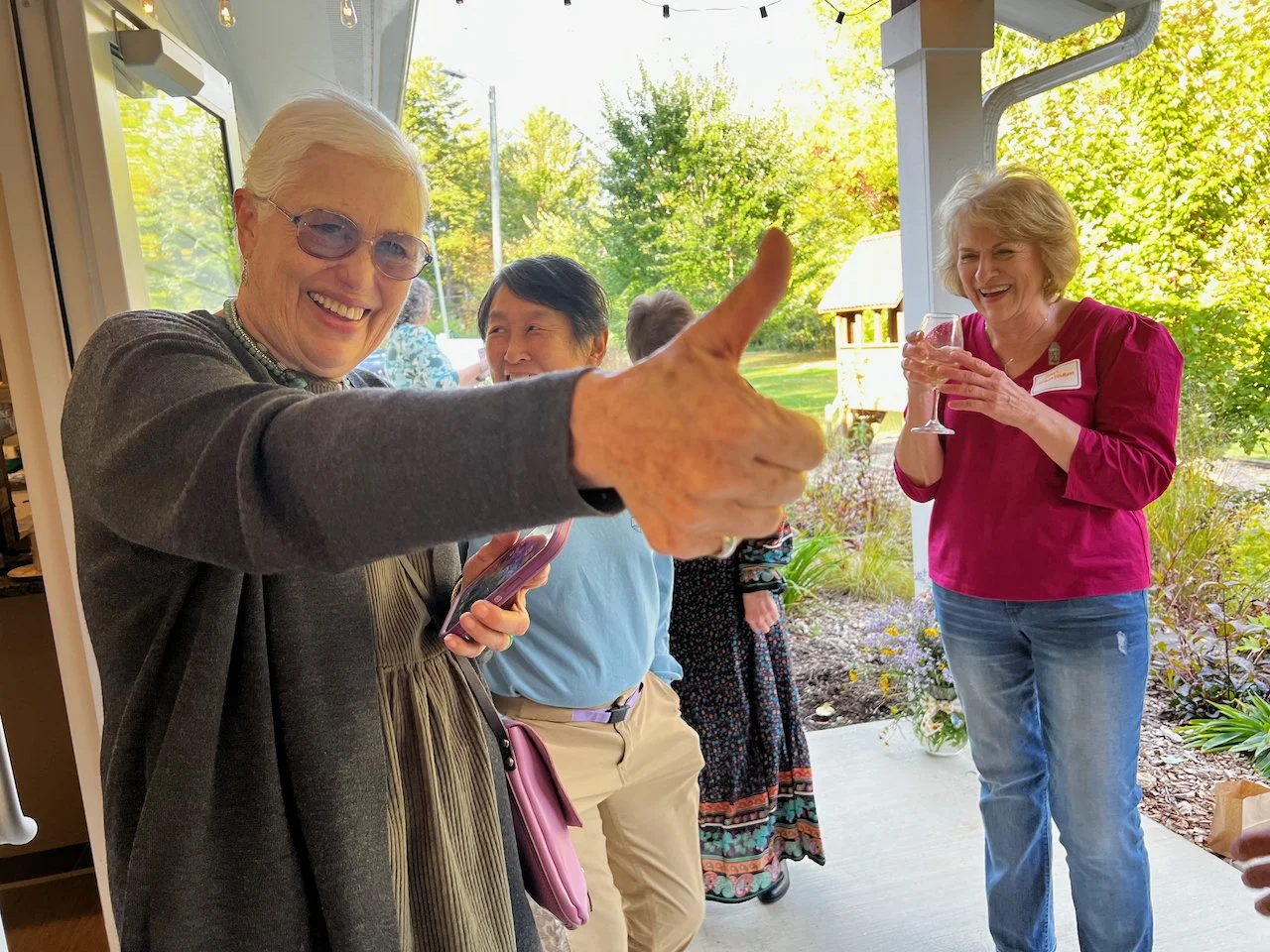 Group of elderly women enjoying socializing outdoors near a porch with trees in the background, one woman giving a thumbs-up.