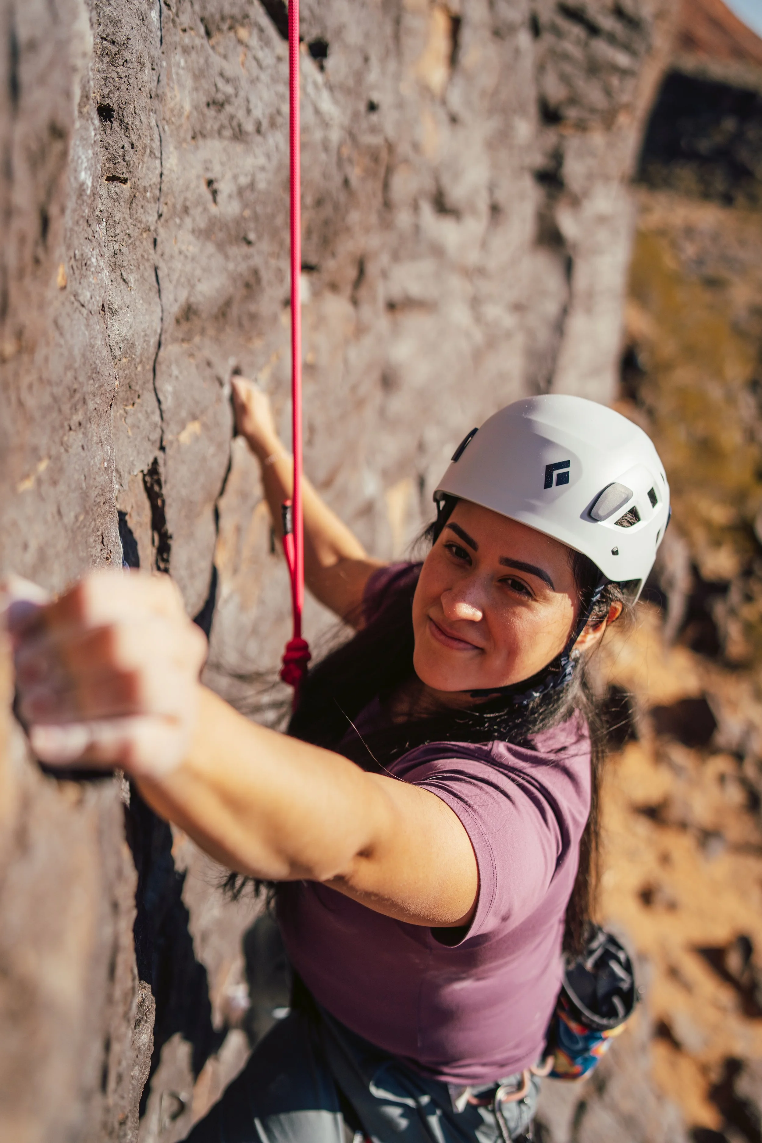 Rock climber wearing a white helmet ascends a rocky cliff using a red climbing rope.
