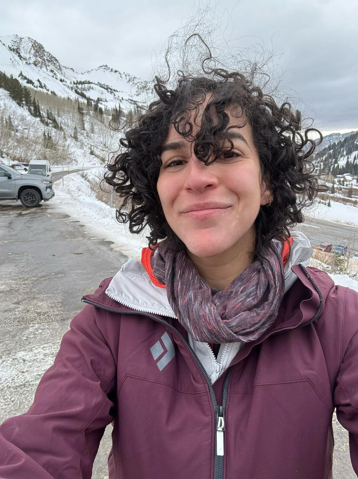 Smiling person in a burgundy winter jacket and scarf taking a selfie in front of snowy mountain slopes.