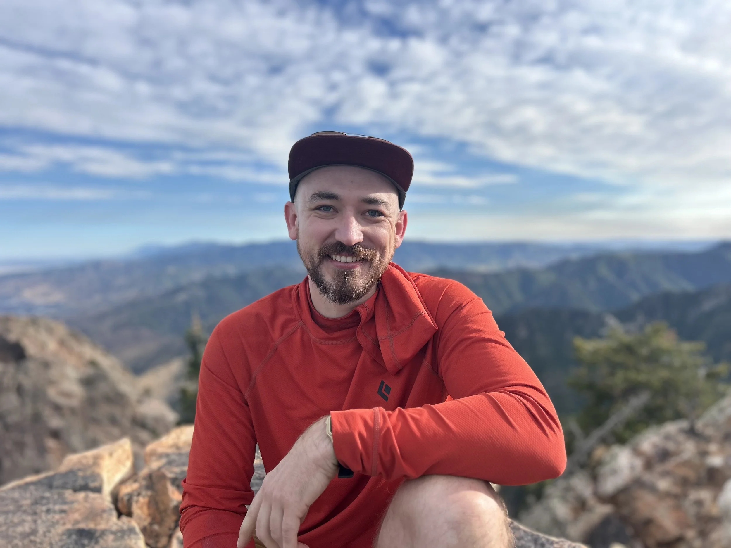 Smiling bearded man in a red hiking shirt and cap taking a selfie on a rocky mountain overlook
