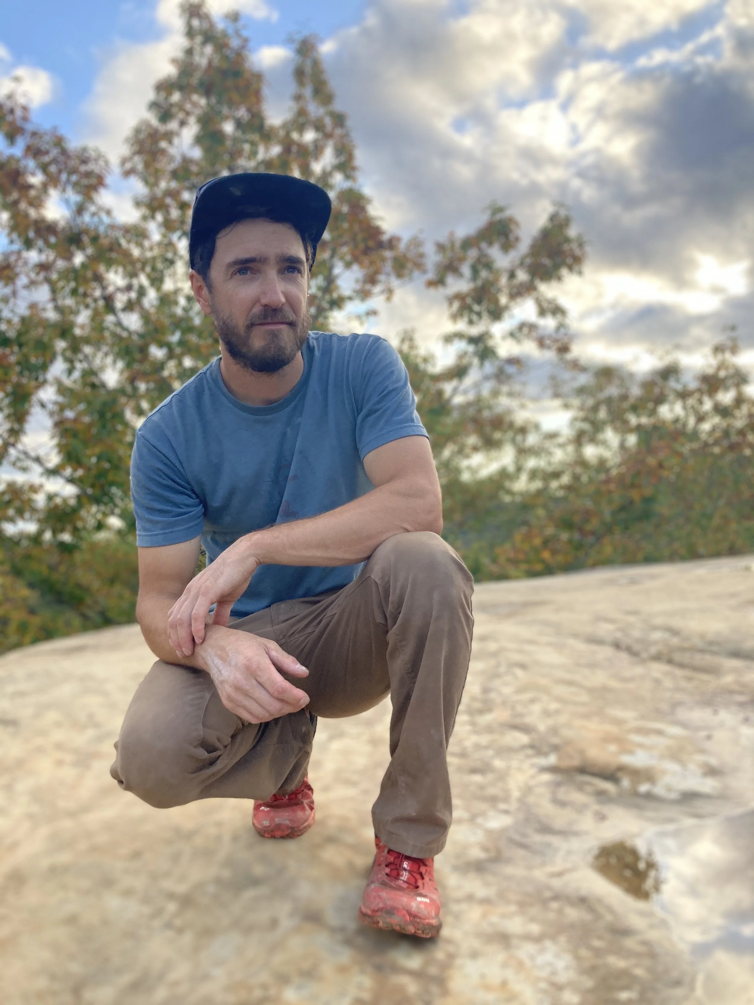 Bearded man in a black cap and blue shirt crouching outdoors beneath a cloudy sky