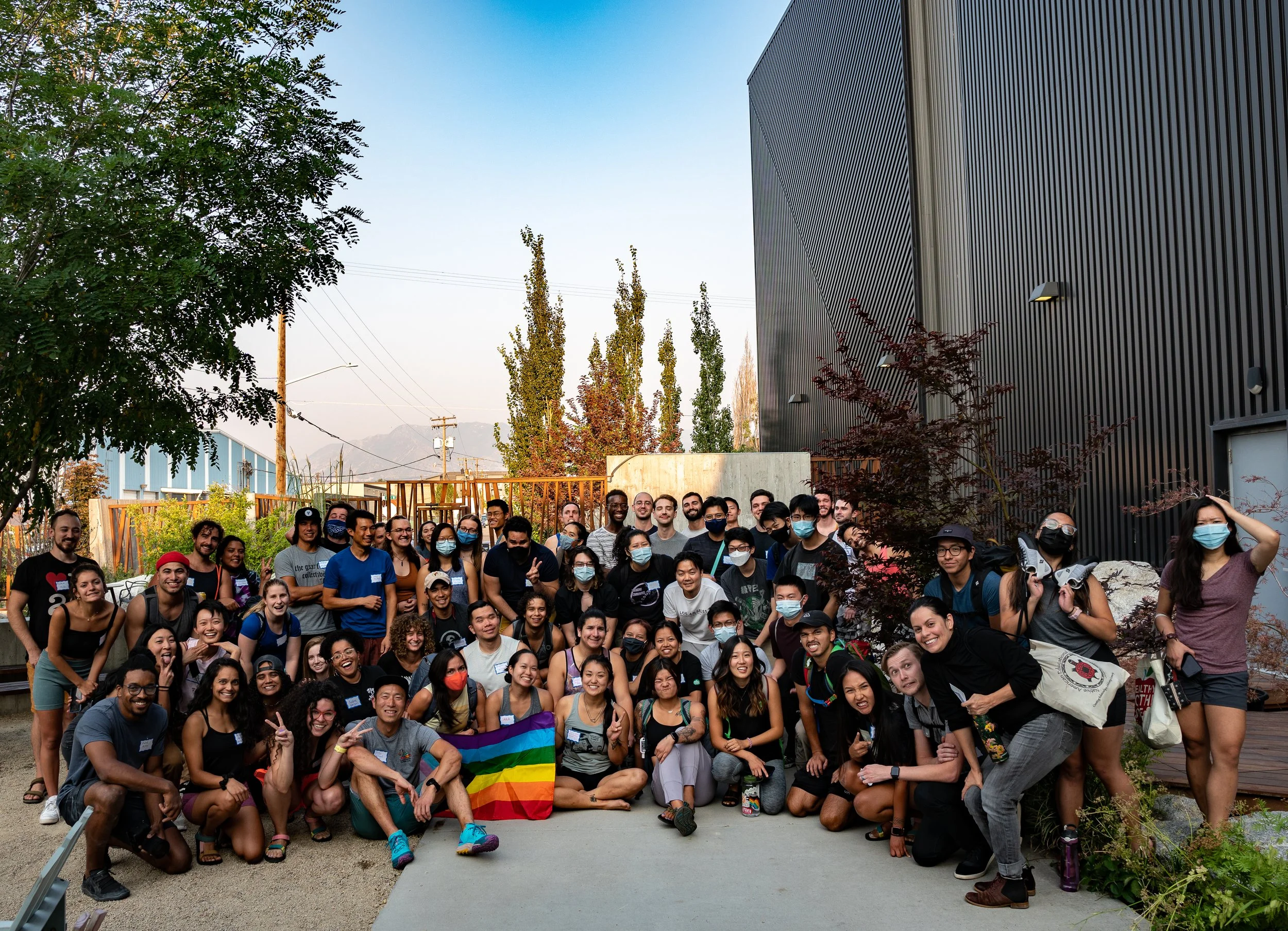 Large diverse group posing outdoors beside a modern industrial building, with trees, mountains, and a rainbow pride flag.