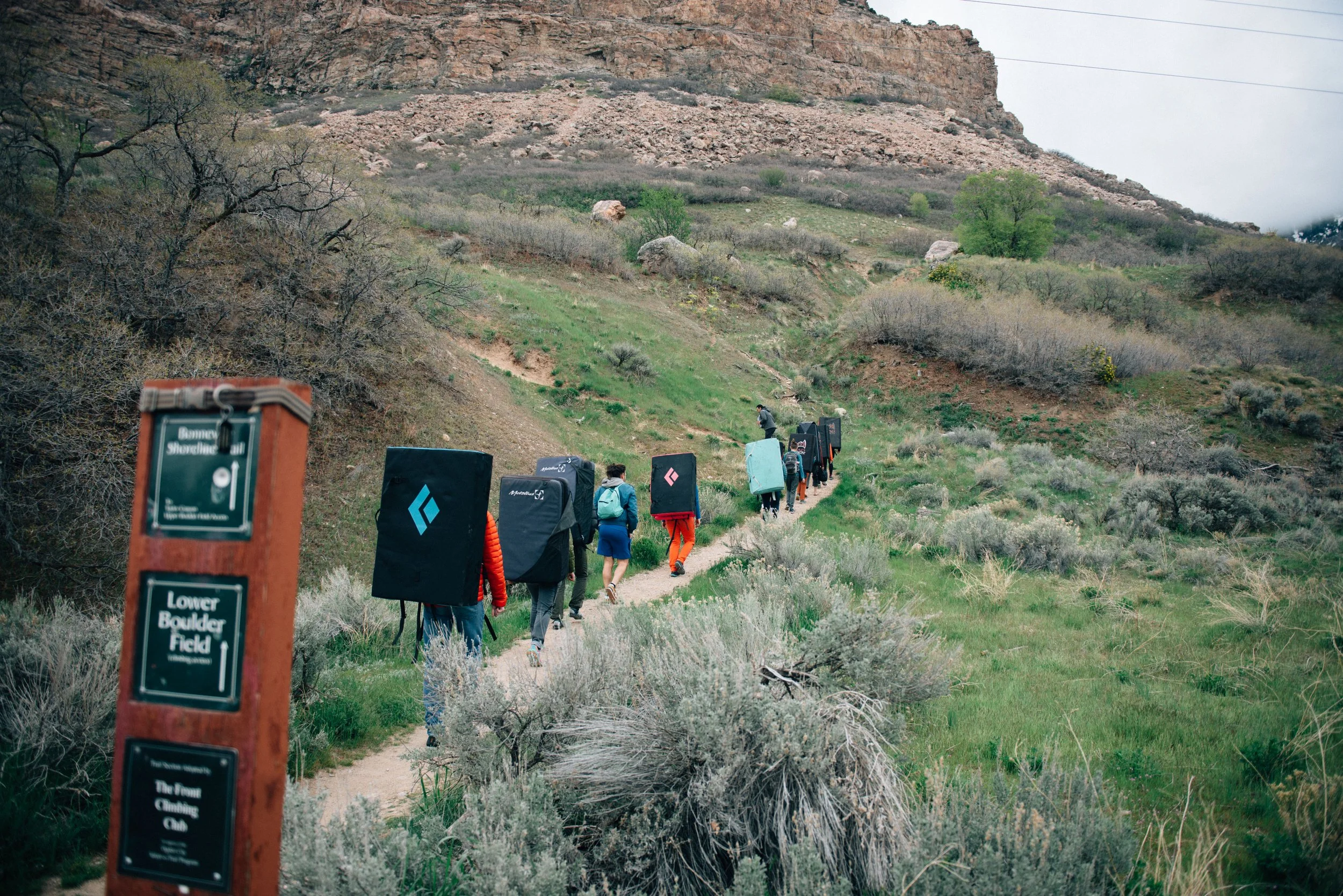 Rock climbers carrying crash pads along a trail beneath a rocky cliff, with a bouldering area sign in the foreground.