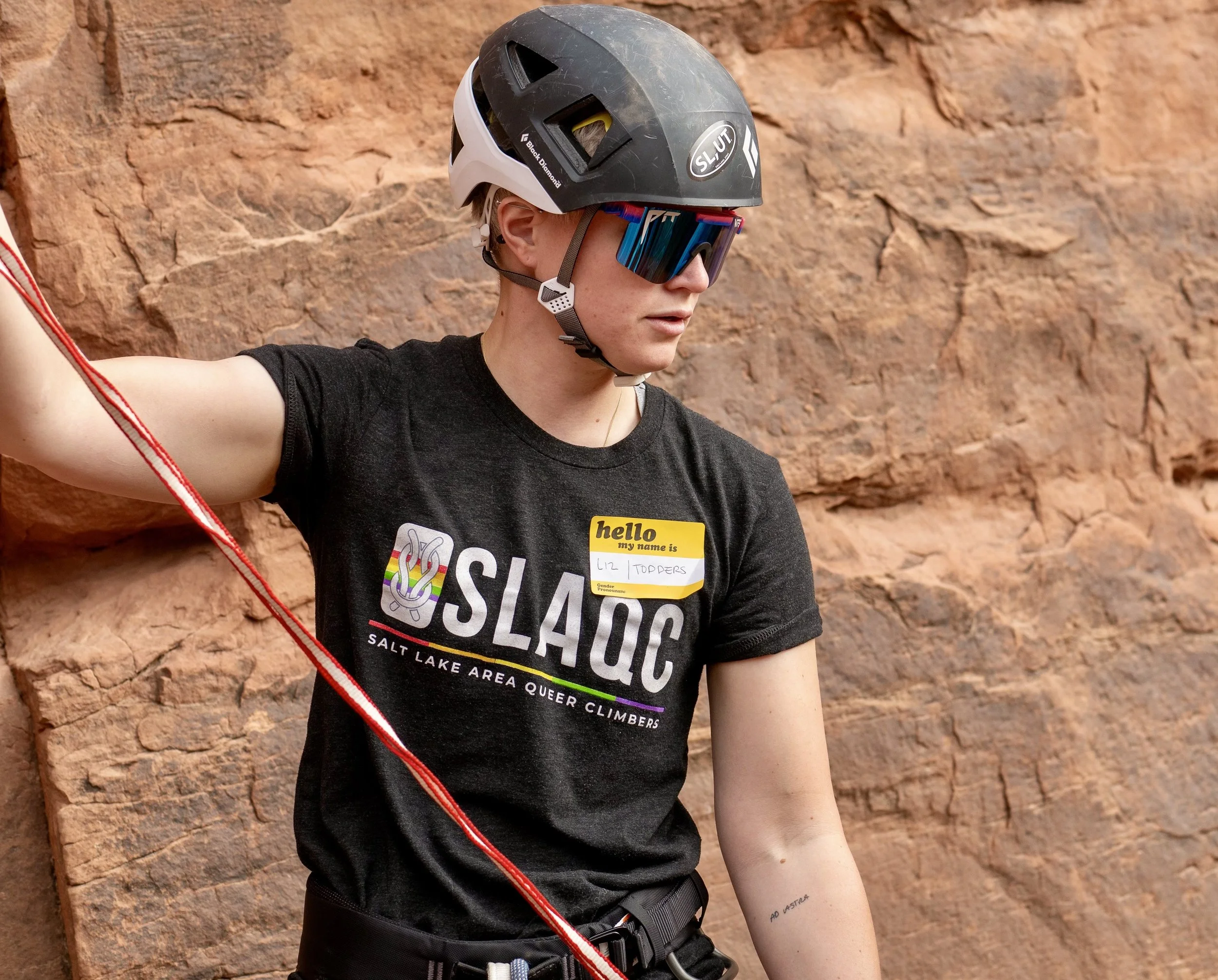 Rock climber wearing a helmet and sunglasses, secured by a red climbing rope against a sandstone wall.