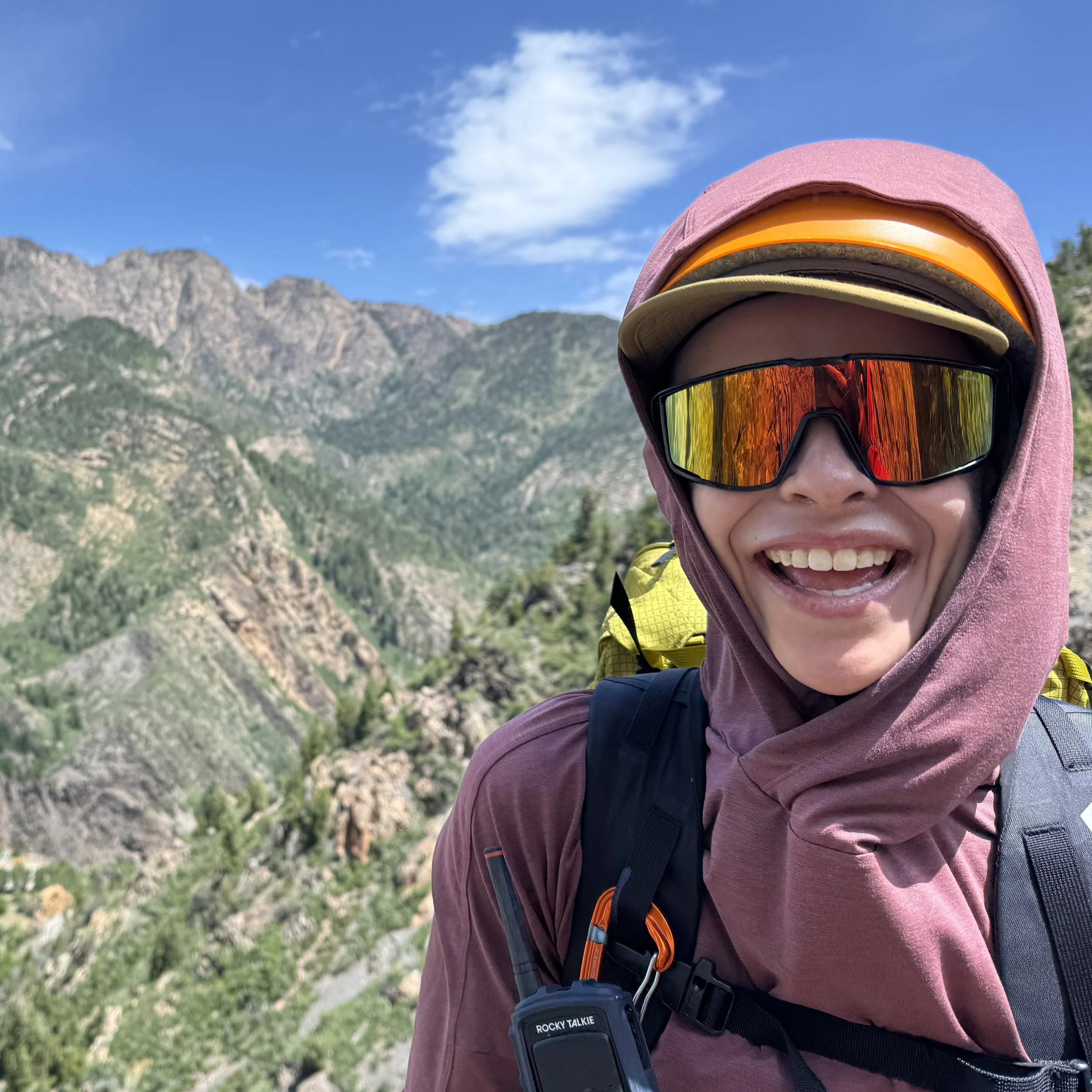 Smiling hiker wearing a hood, pink cap, and reflective sunglasses with rugged mountains behind her