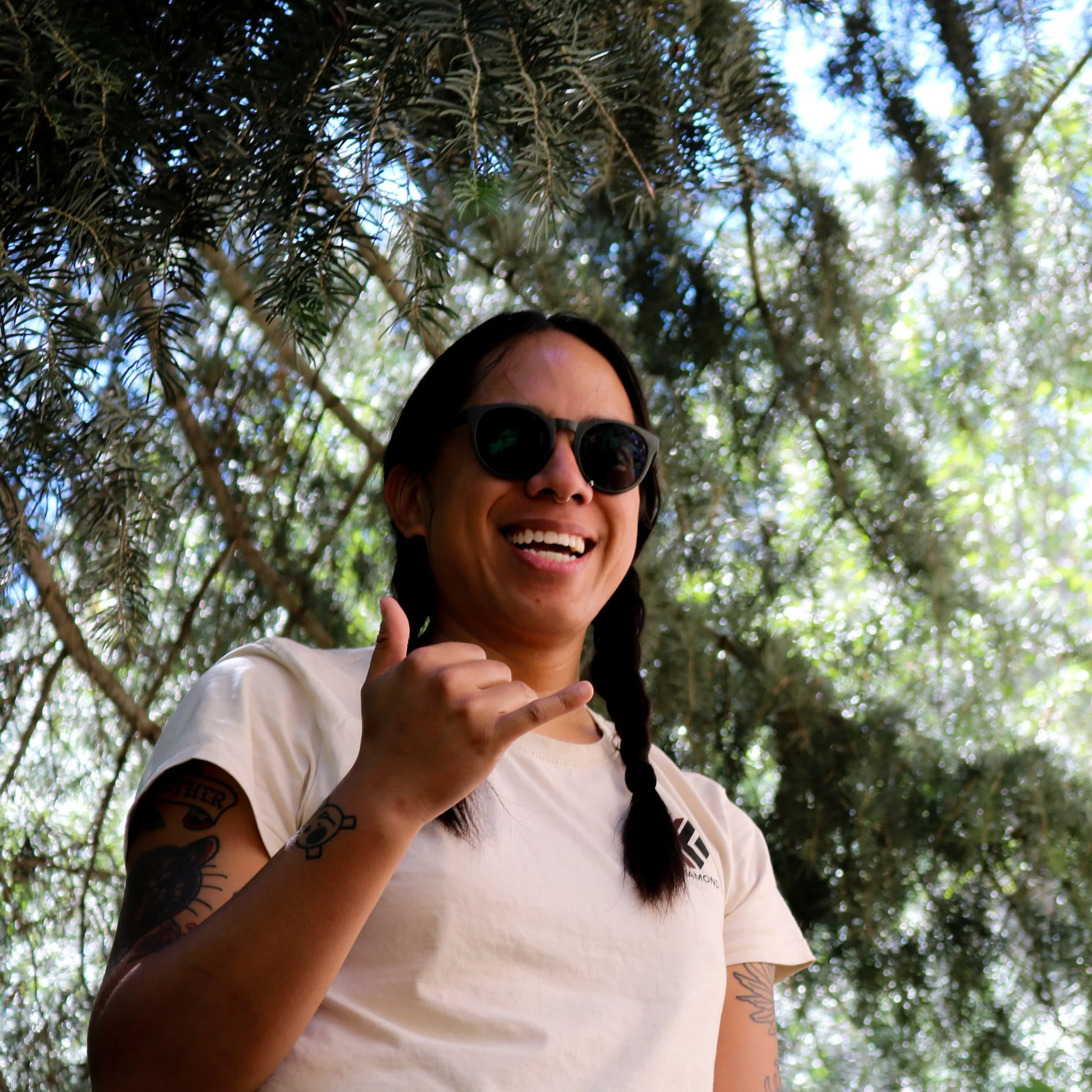 Smiling tattooed woman wearing sunglasses gives a shaka hand gesture outdoors beneath leafy trees
