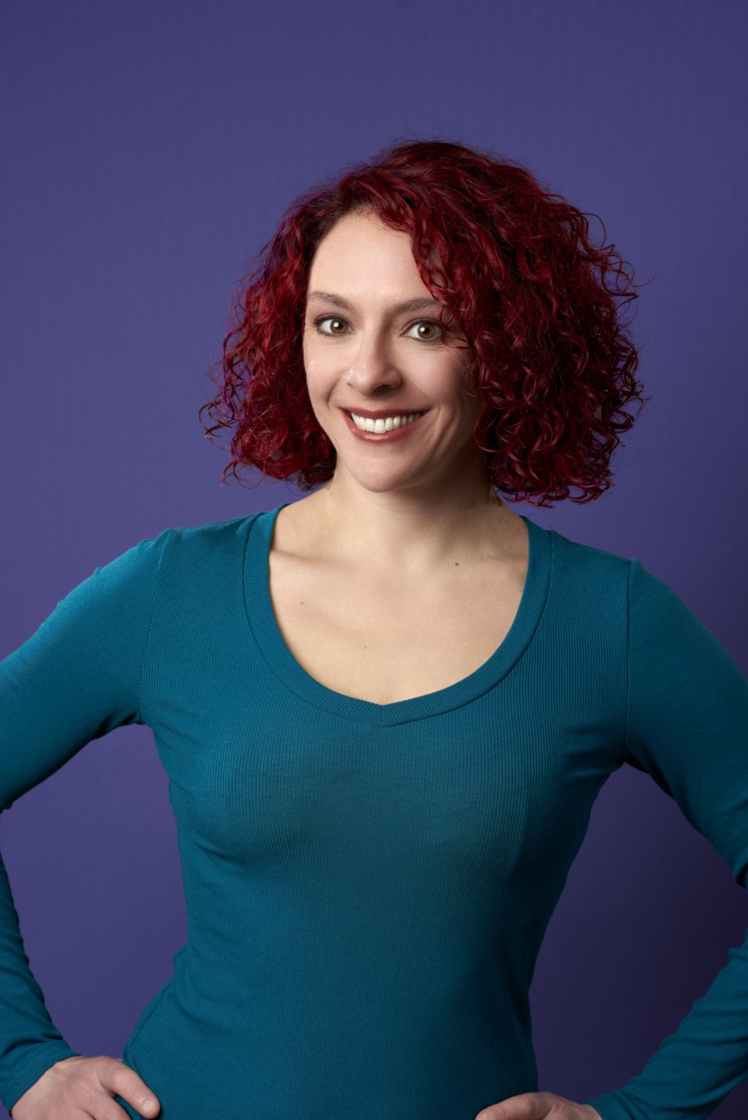 Portrait of a woman with curly red hair, wearing a long sleeve teal shirt, smiling against a purple background.