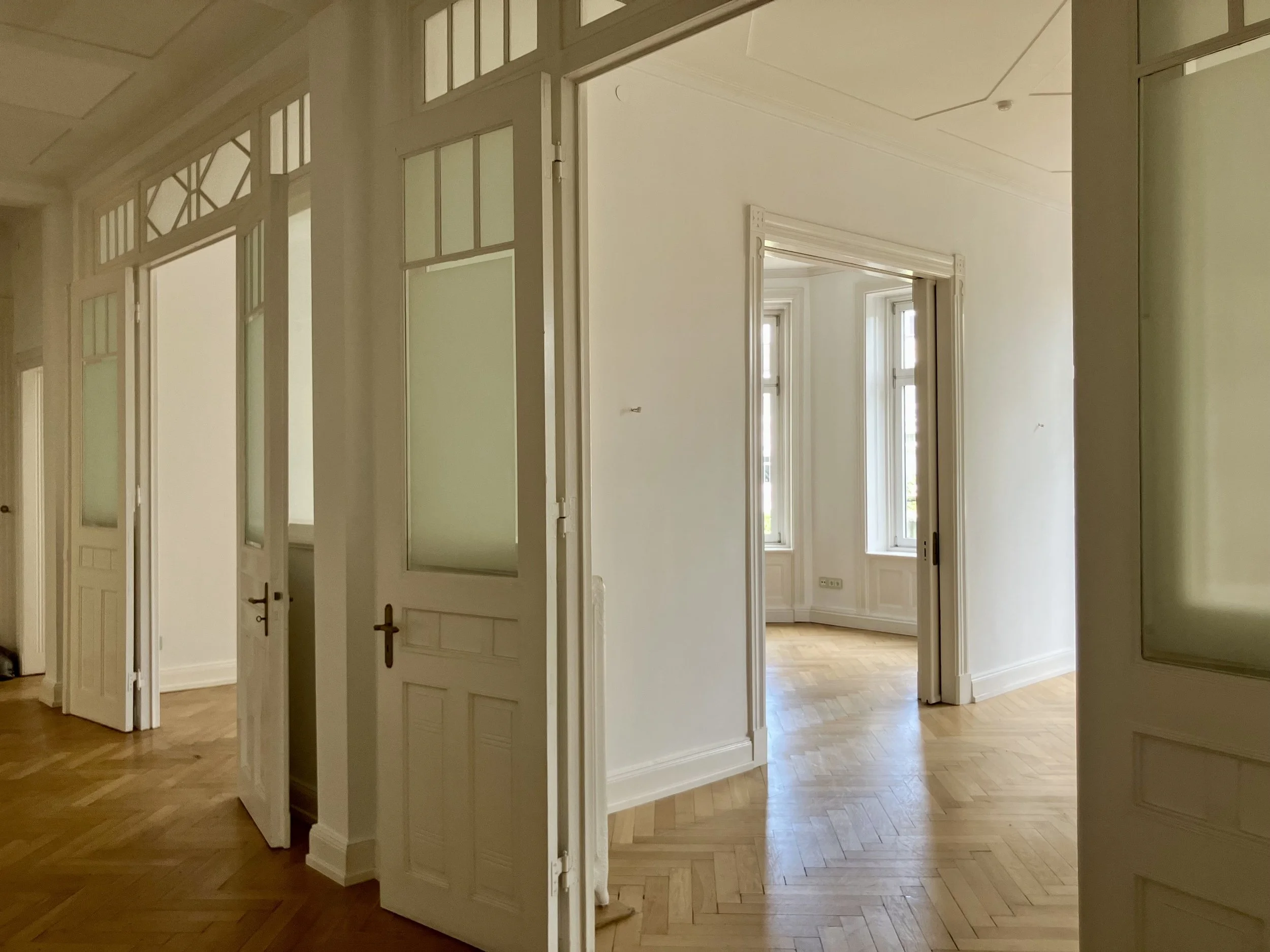 A hallway in Vienna with multiple white wooden doors featuring glass panels, open to reveal rooms with large windows letting in natural light. The floor is wooden parquet, and the walls are painted white.