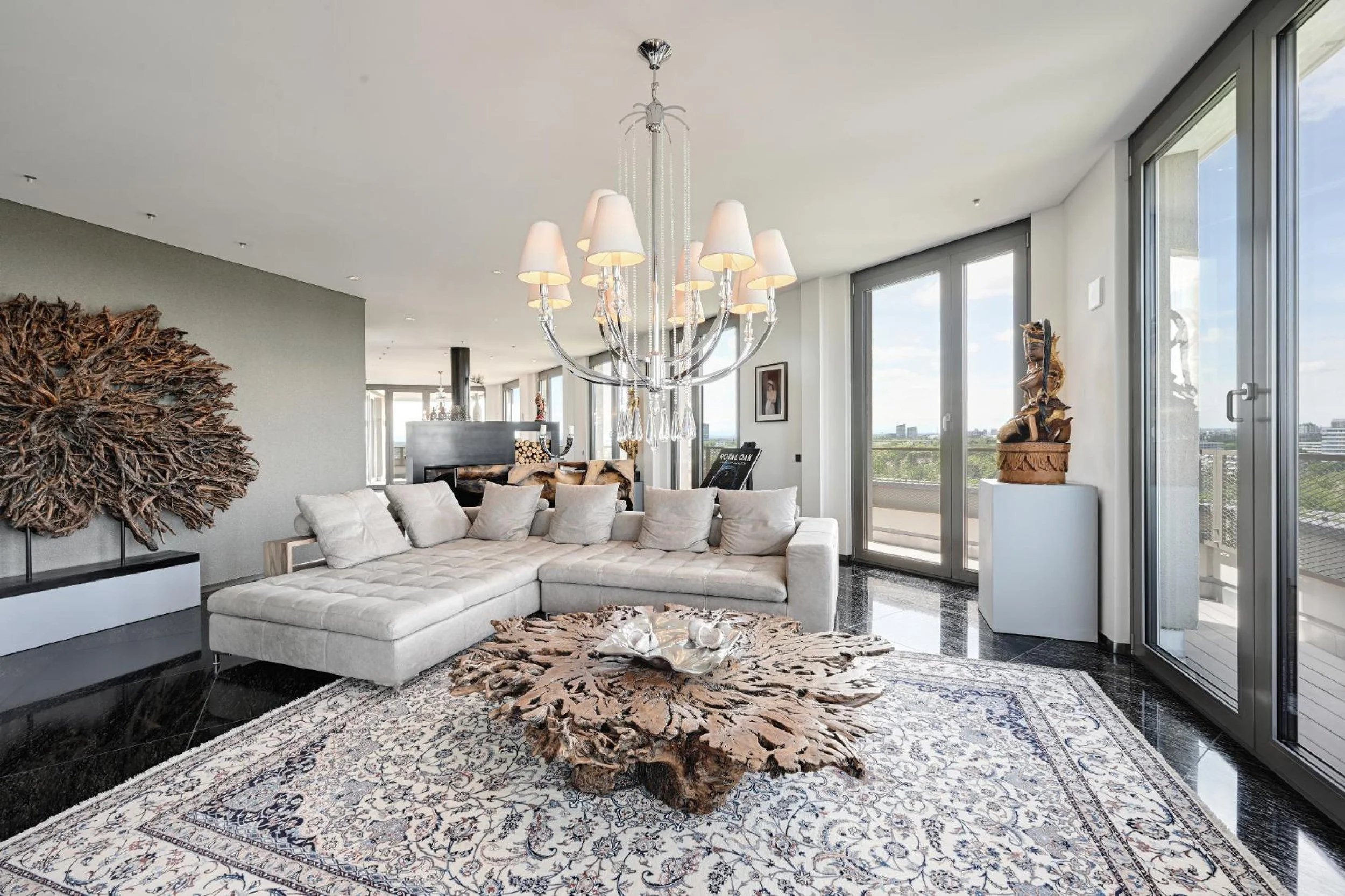 Modern Munich living room with a large L-shaped beige sofa, a wooden coffee table, ornate area rug, a white wall with a textured art piece, a chandelier, large windows with a city view, and sculptures on white pedestals.