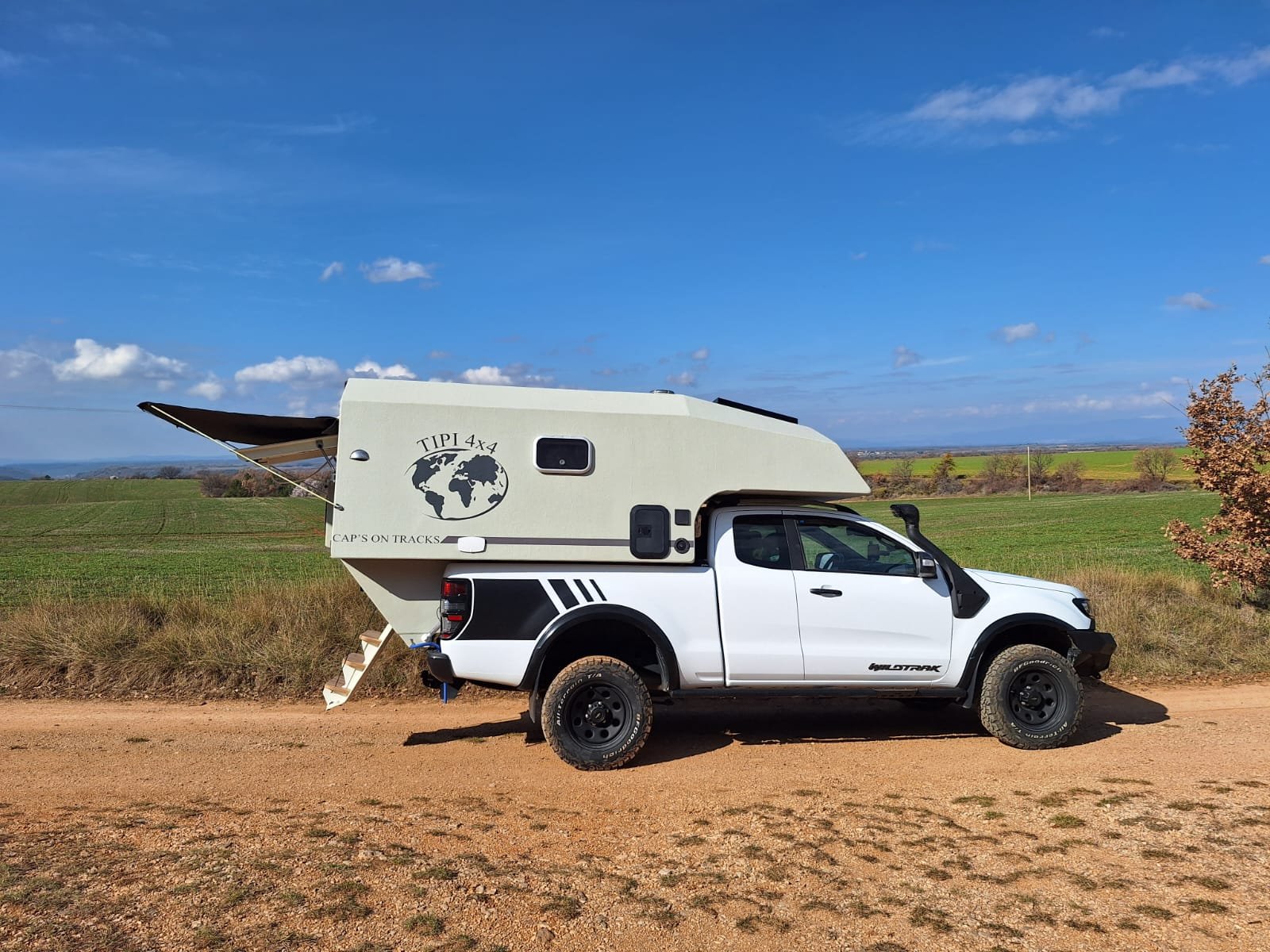 Camionnette blanche avec cellule de camping "Tipi 4x4" sur piste de terre, paysage champêtre en arrière-plan.