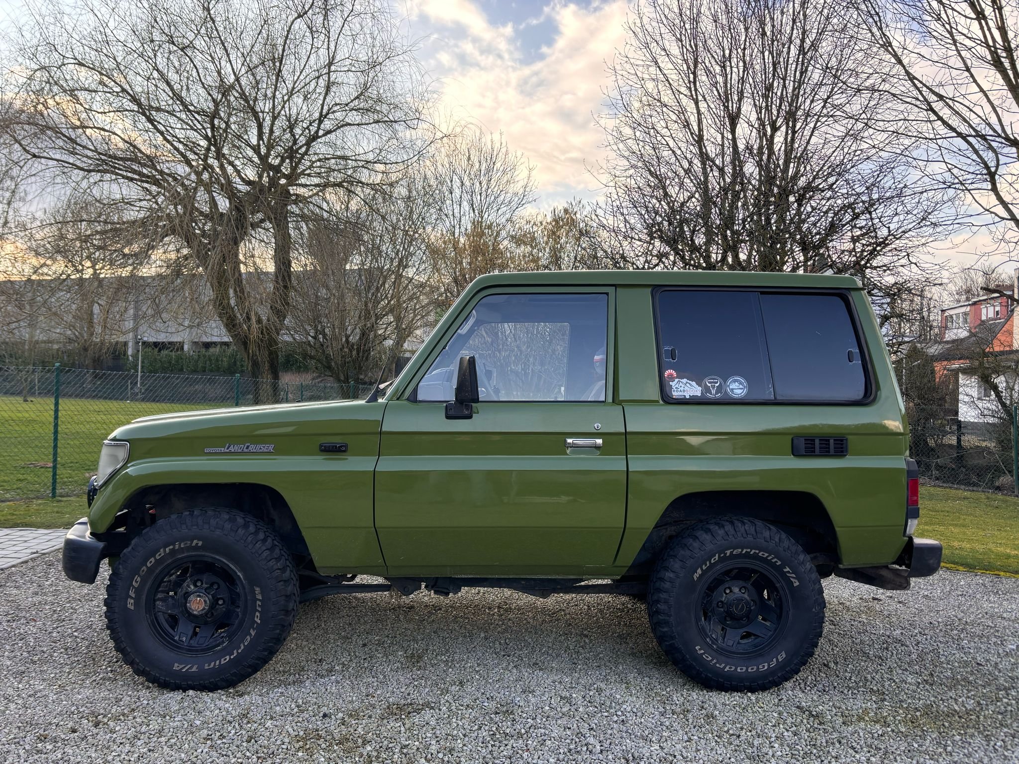 Toyota Land Cruiser vert stationné sur un chemin gravillonné, avec des arbres dénudés à l'arrière-plan.