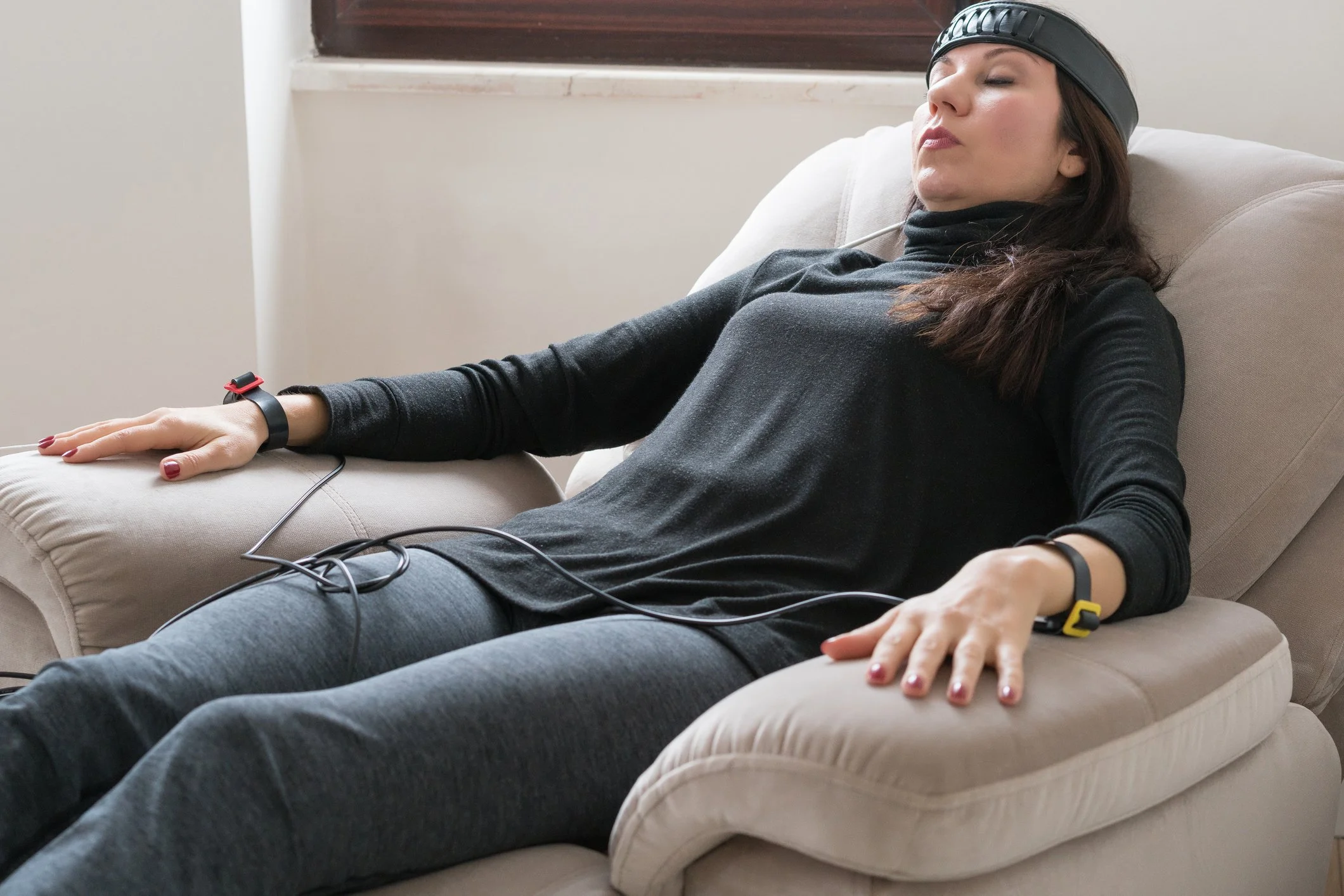 Woman relaxing on a beige chair with her eyes closed, wearing a black headband and casual clothing, with electrodes attached to her head and wrist, connected to wires.