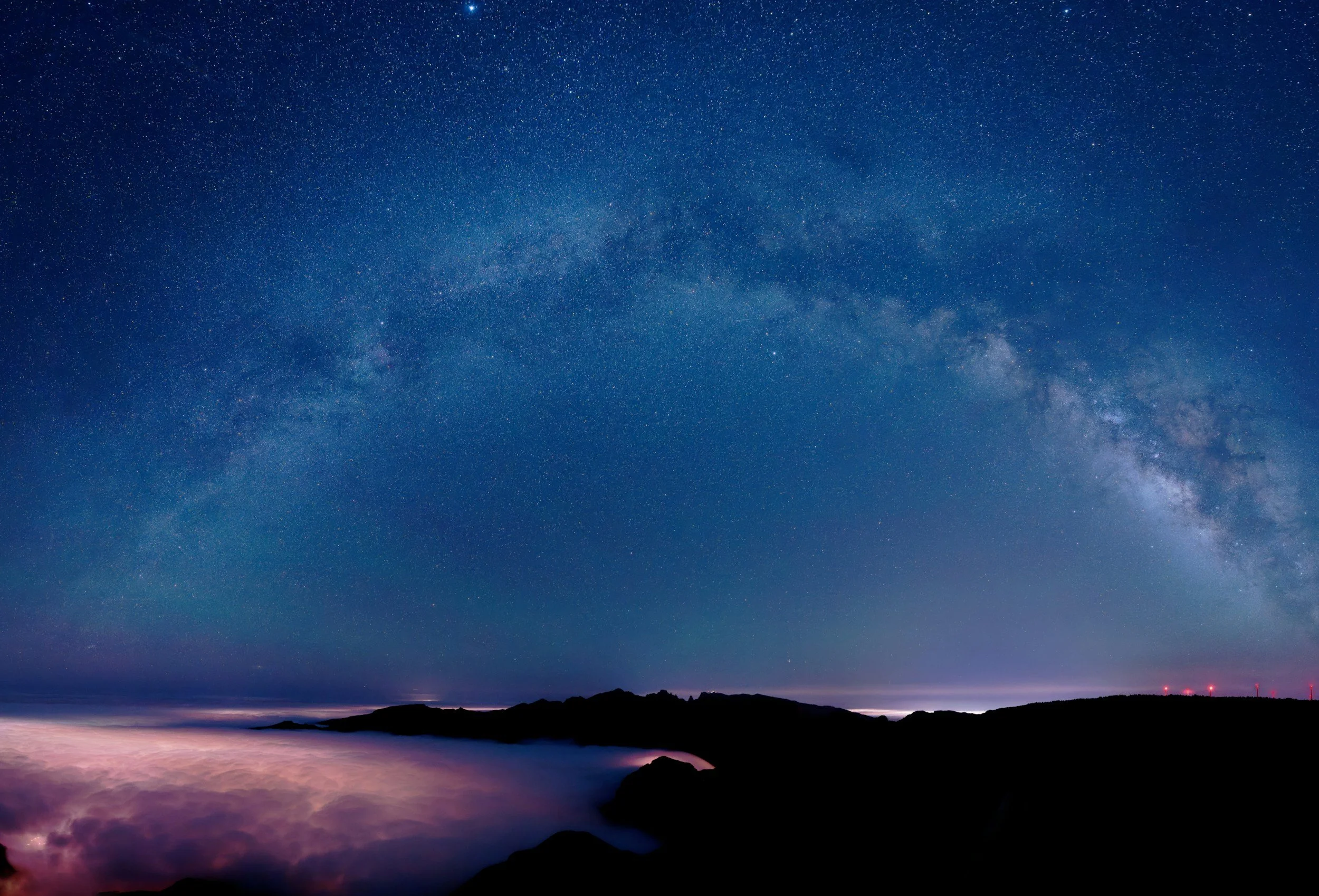 Nighttime landscape with a star-filled sky and the Milky Way galaxy arcing overhead, above a dark coastline with rocks and fog or clouds near the water.