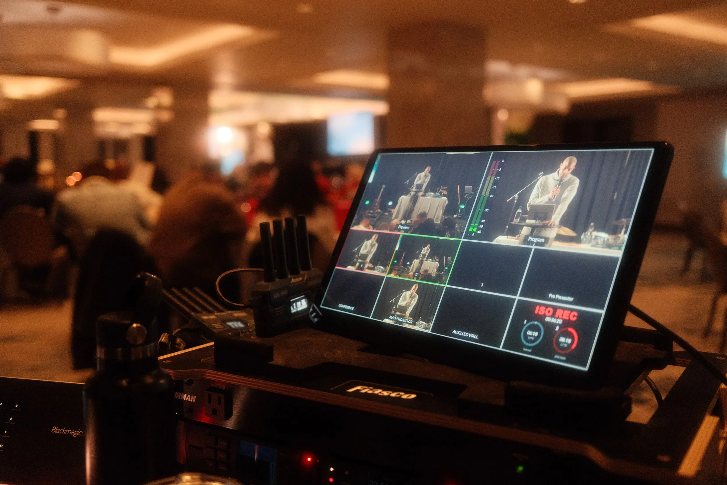 View of an event setup indoors with a camera and a monitor in the foreground, show chairs arranged for an audience, tables with people dining, a stage area with cameras and equipment, and a screened area with attendees, in a modern venue with glass walls and ceiling.