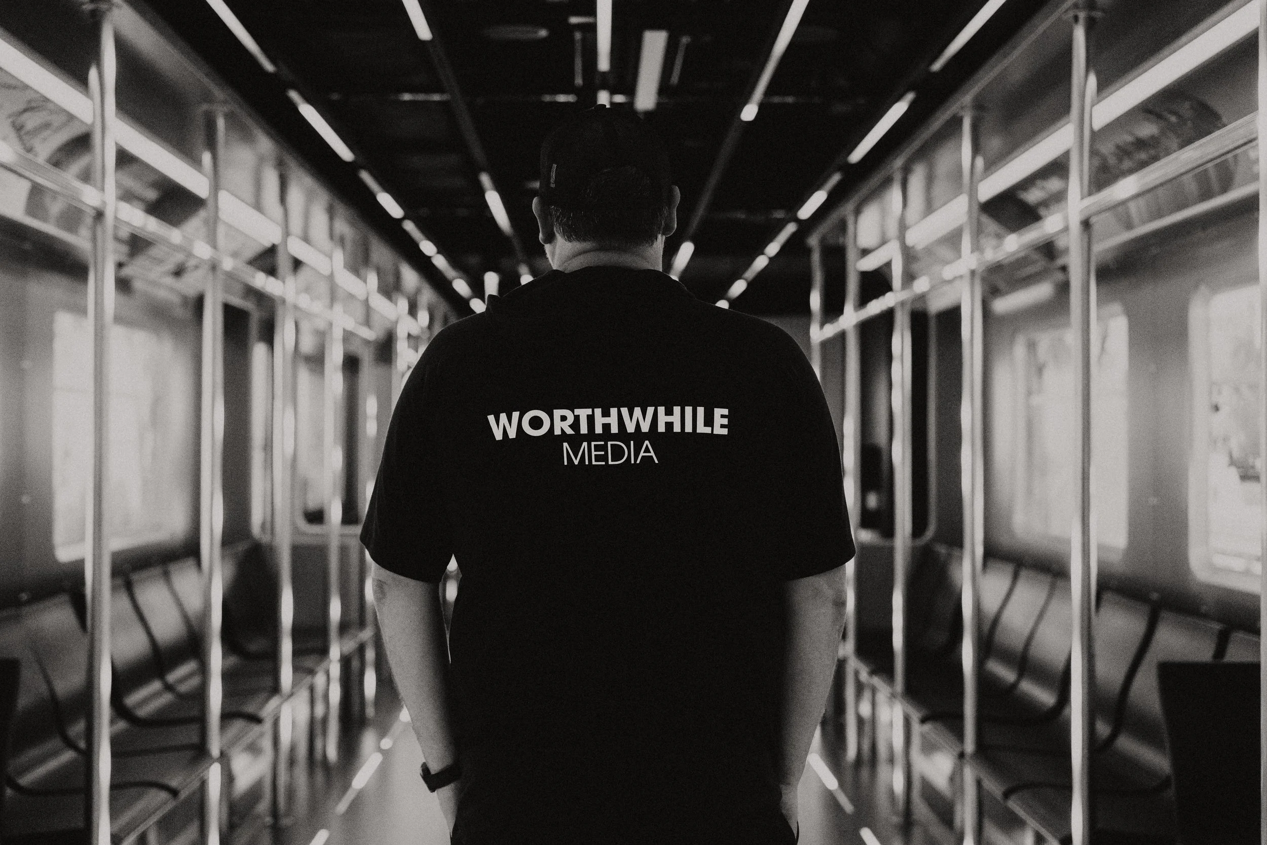 A man wearing a black shirt with the words "WORTHWHILE MEDIA" on the back, standing in an empty black and white subway train car, facing away from the camera.