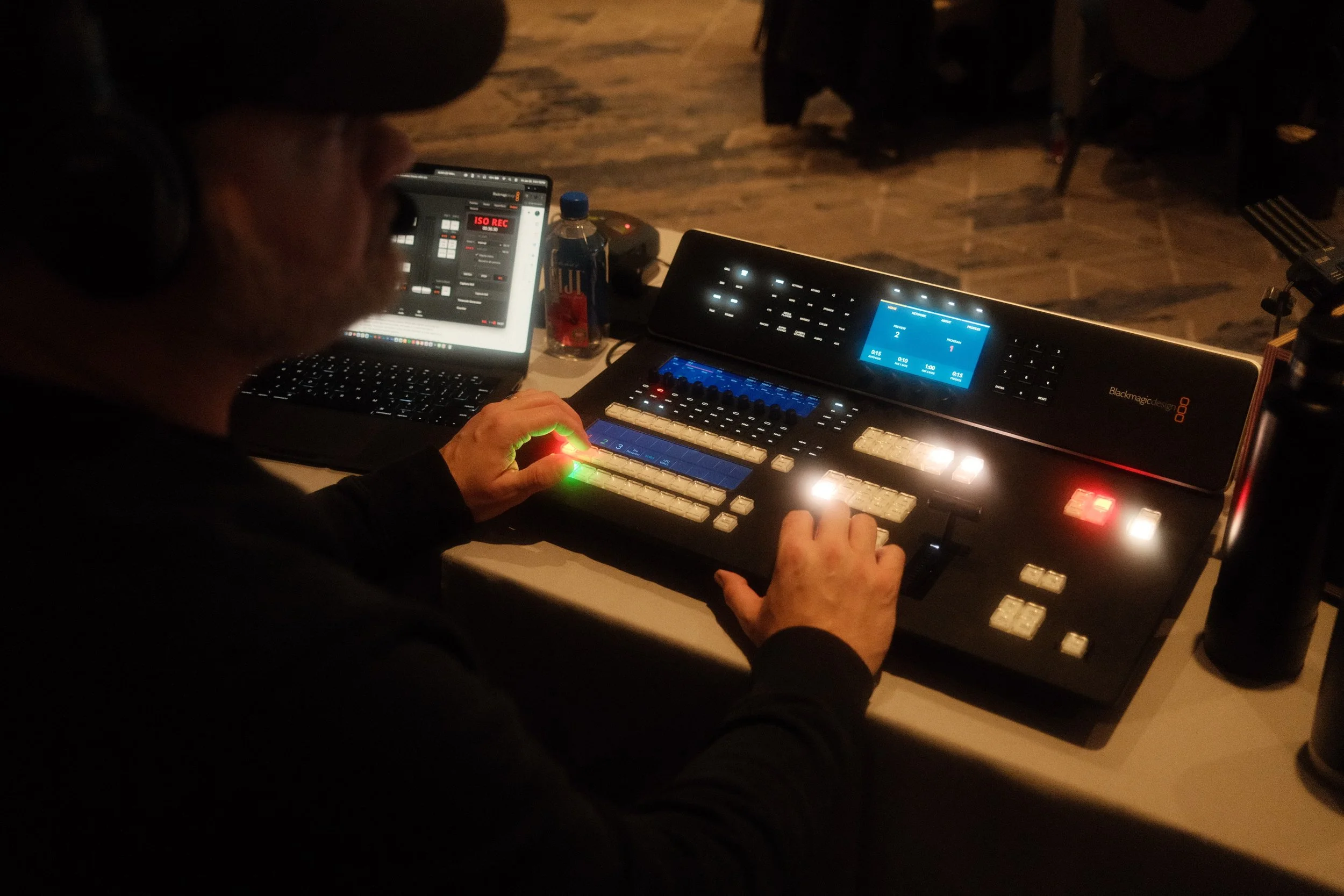 A black and white photo of an interview or conversation being recorded indoors. A woman on the left holds a phone, facing two people on the right seated on a couch, one woman and one man. There is a large studio light overhead, and the background wall has words such as 'Spirit Empowered' and 'Cultural'.