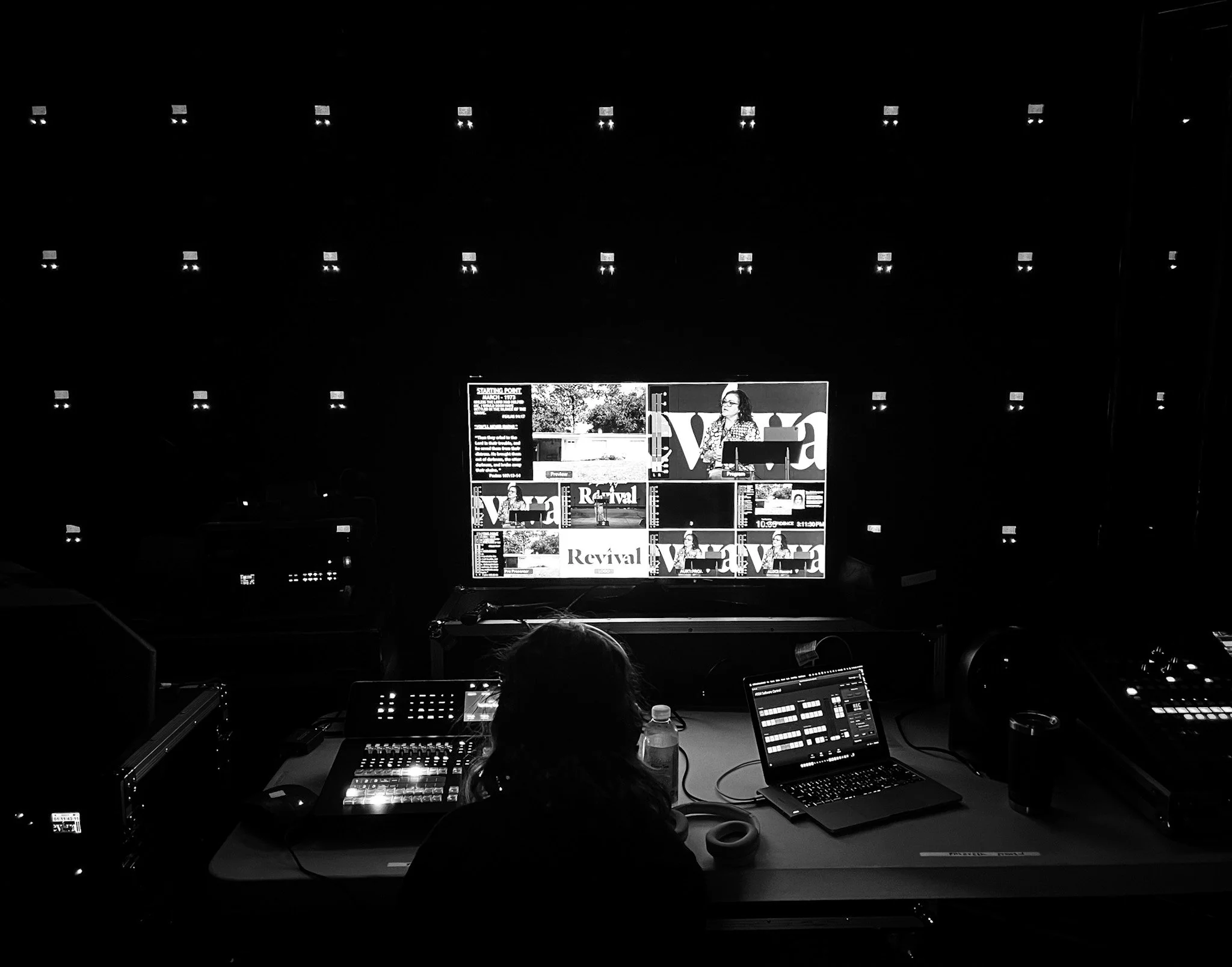 A person working at a control station with a laptop, audio equipment, and a water bottle, facing a large screen showing a digital presentation or broadcast in a dark room.