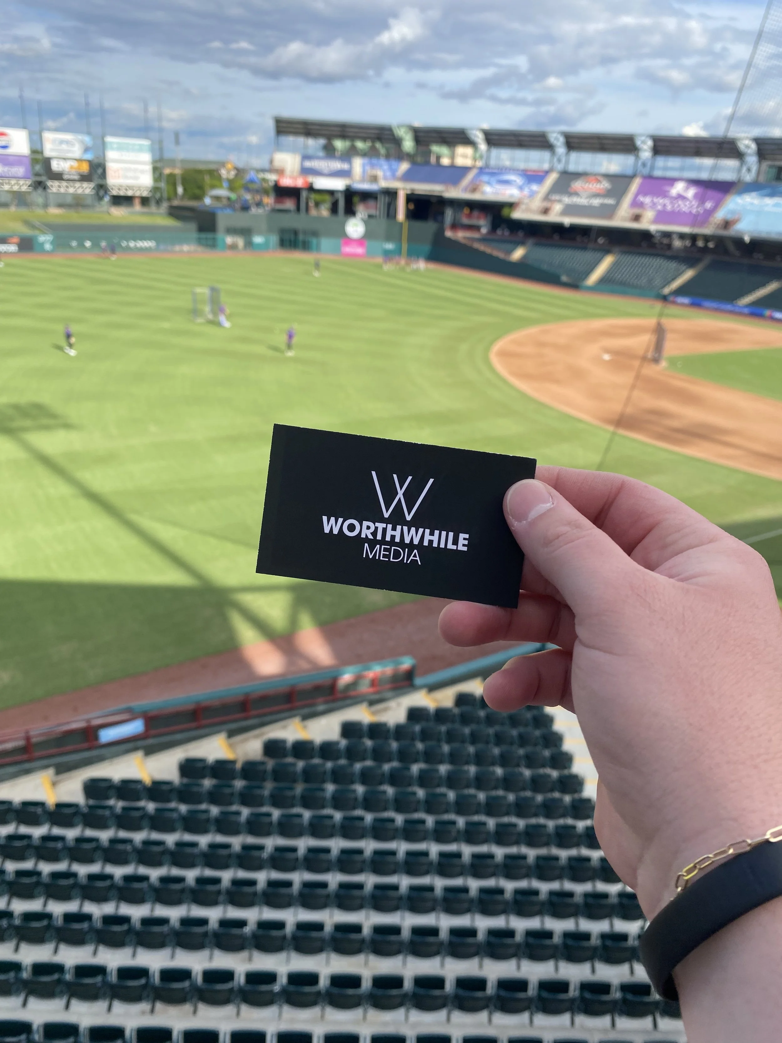 A person holding a black card with the white text 'WORTHWHILE MEDIA' in front of an empty baseball stadium, with players practicing on the field and blue sky with clouds overhead.
