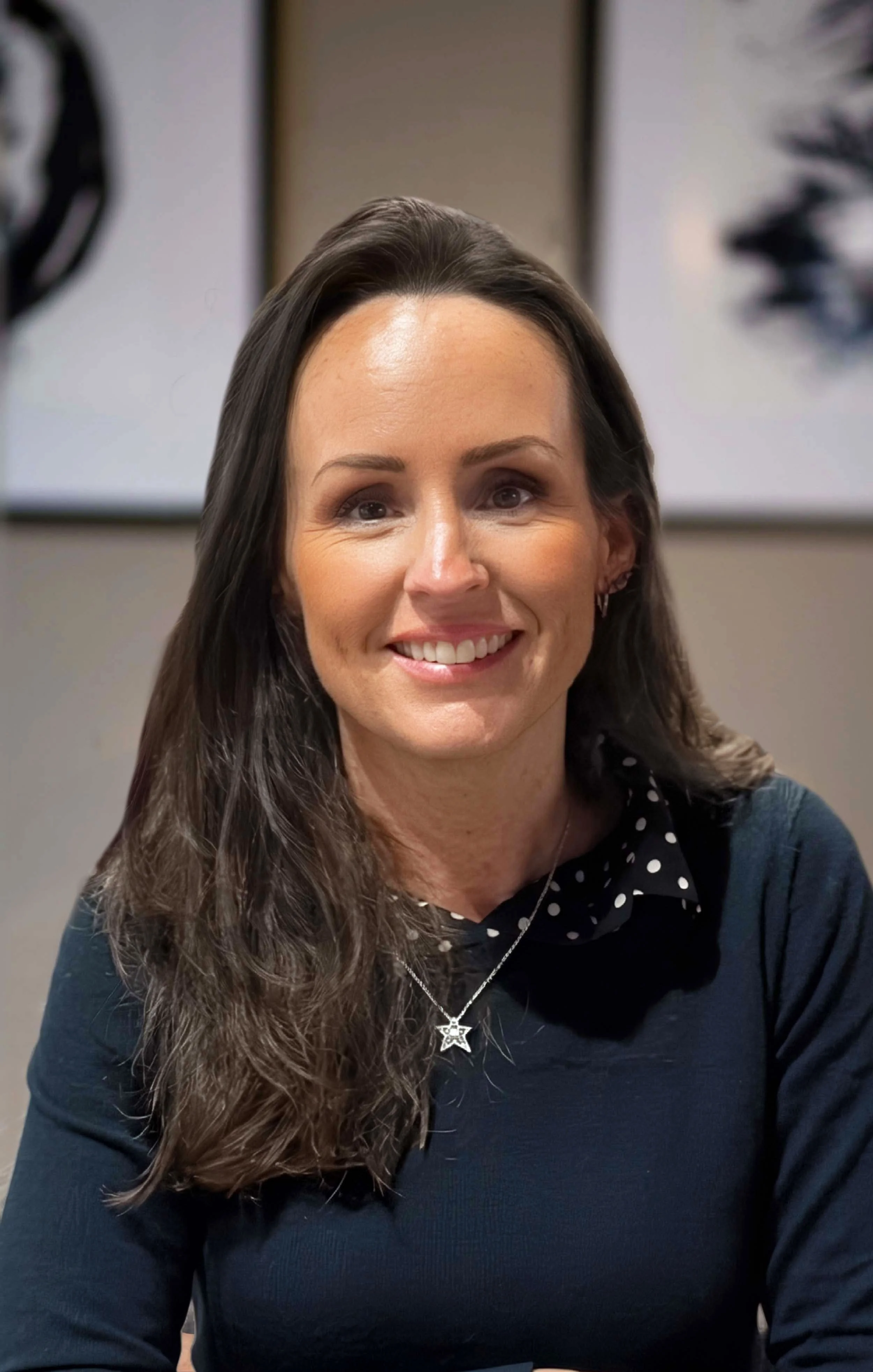 A woman with long, wavy brown hair, wearing earrings, a star-shaped necklace, a polka-dot shirt, and a dark sweater, smiling in front of a blurred framed artwork.