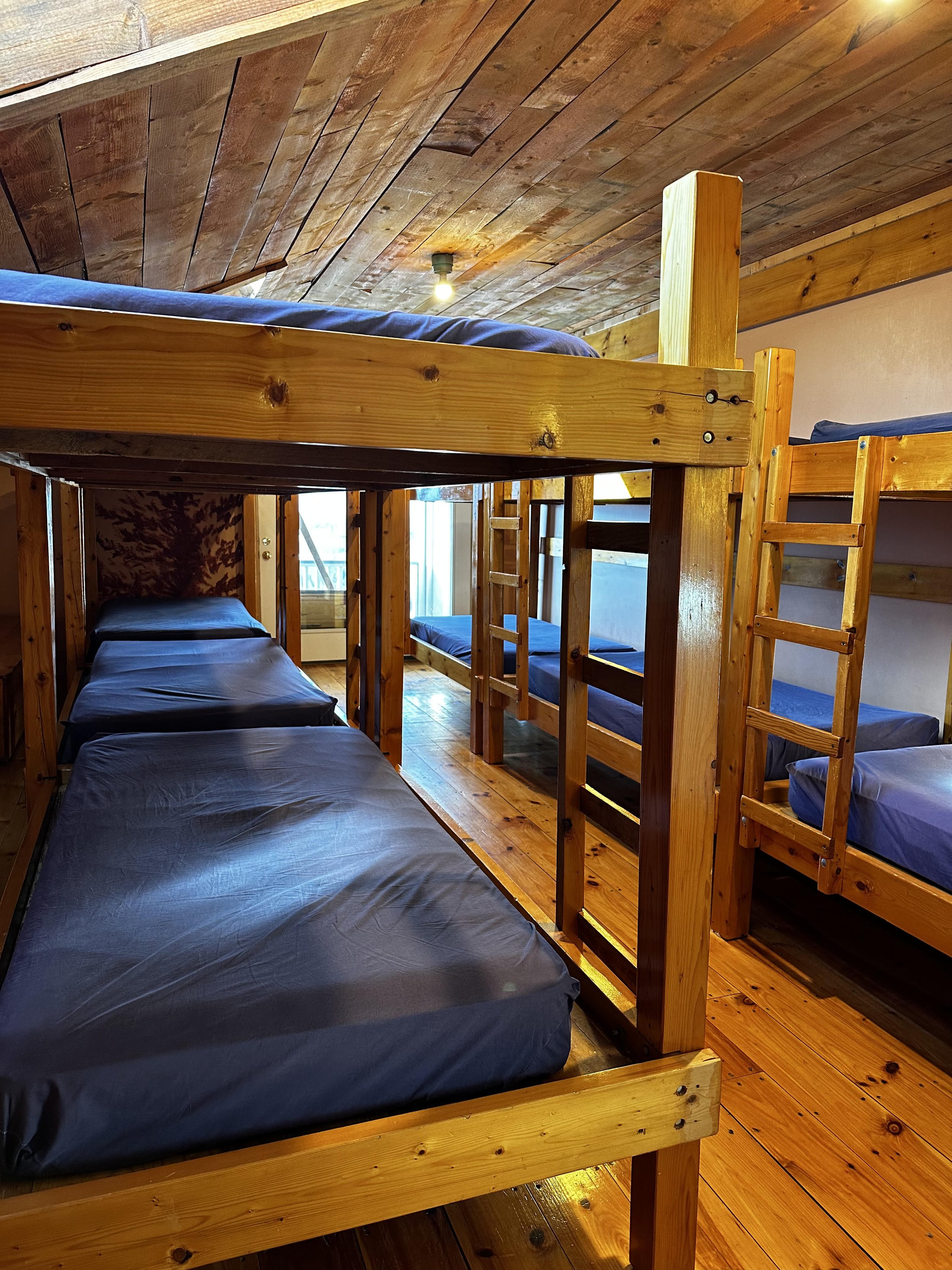 Indoor room with wooden bunk beds, each with a ladder, and blue mattress covers, hardwood floor, and wood-paneled ceiling.