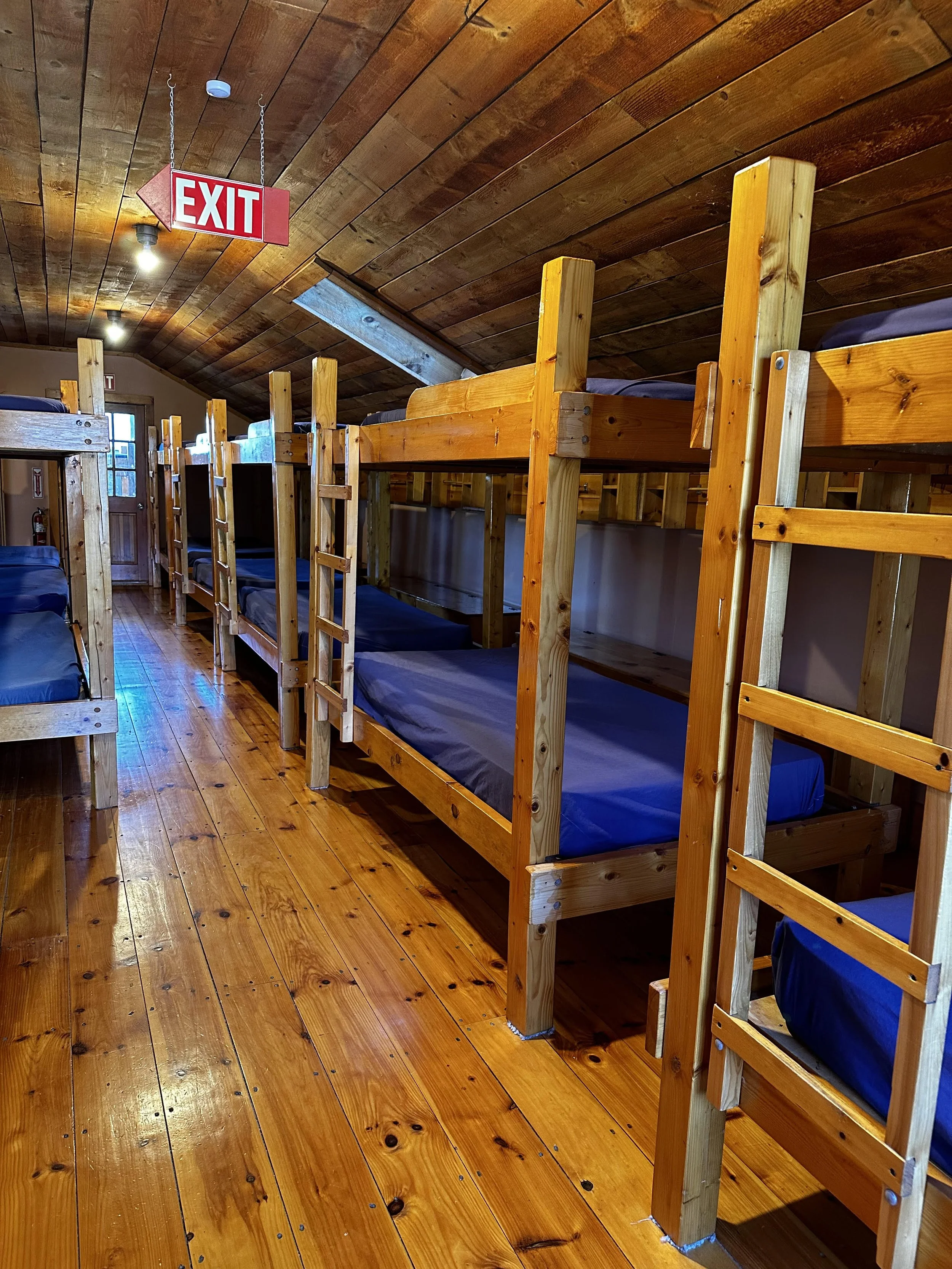 A room with wooden bunk beds and a wooden ceiling, with an exit sign hanging from the ceiling.