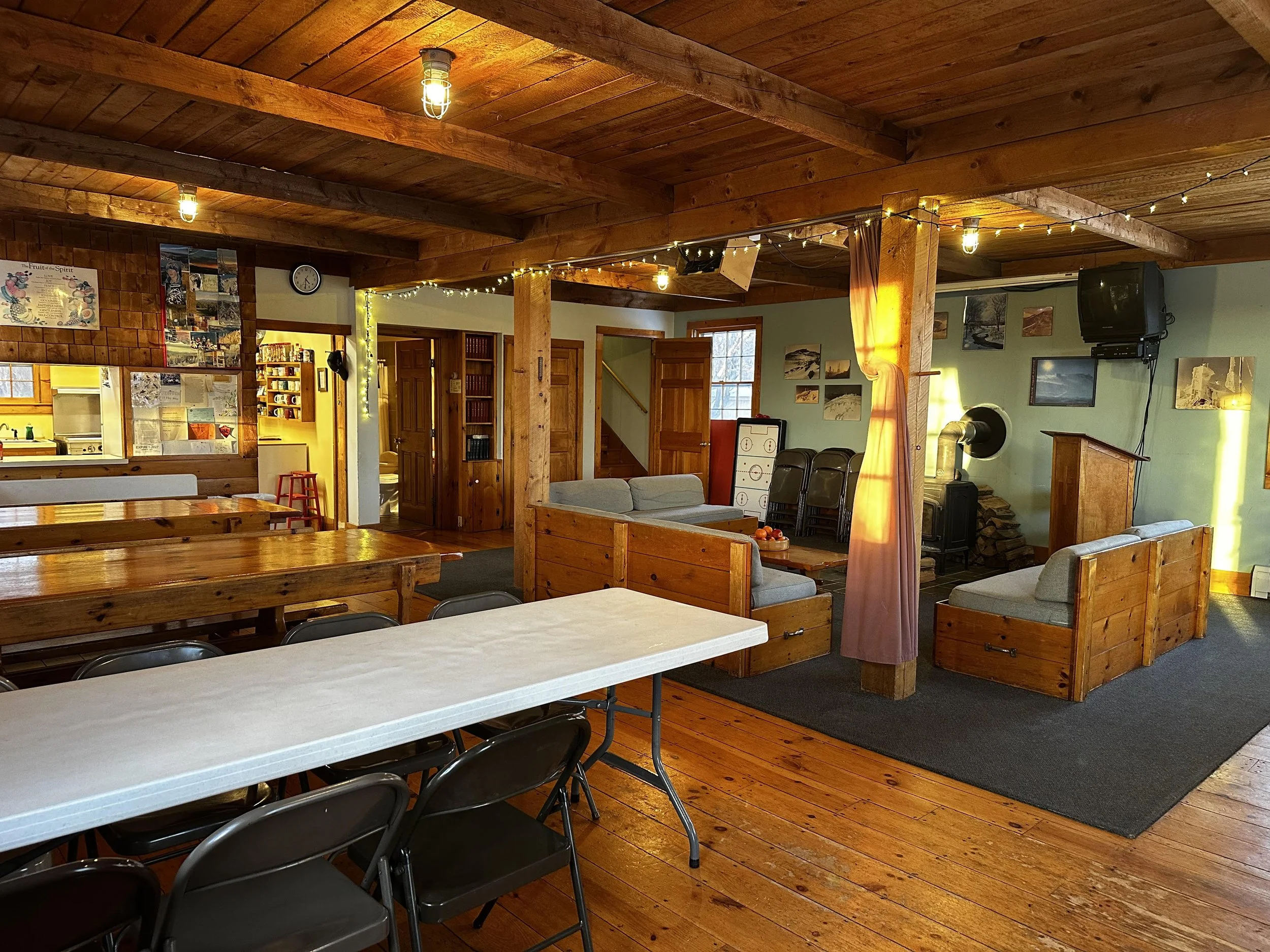 The image shows a cozy, rustic living room with wooden beams and paneling, featuring a seating area with sofas, chairs, and a TV, along with a dining area with a long table and chairs, and a kitchen in the background.