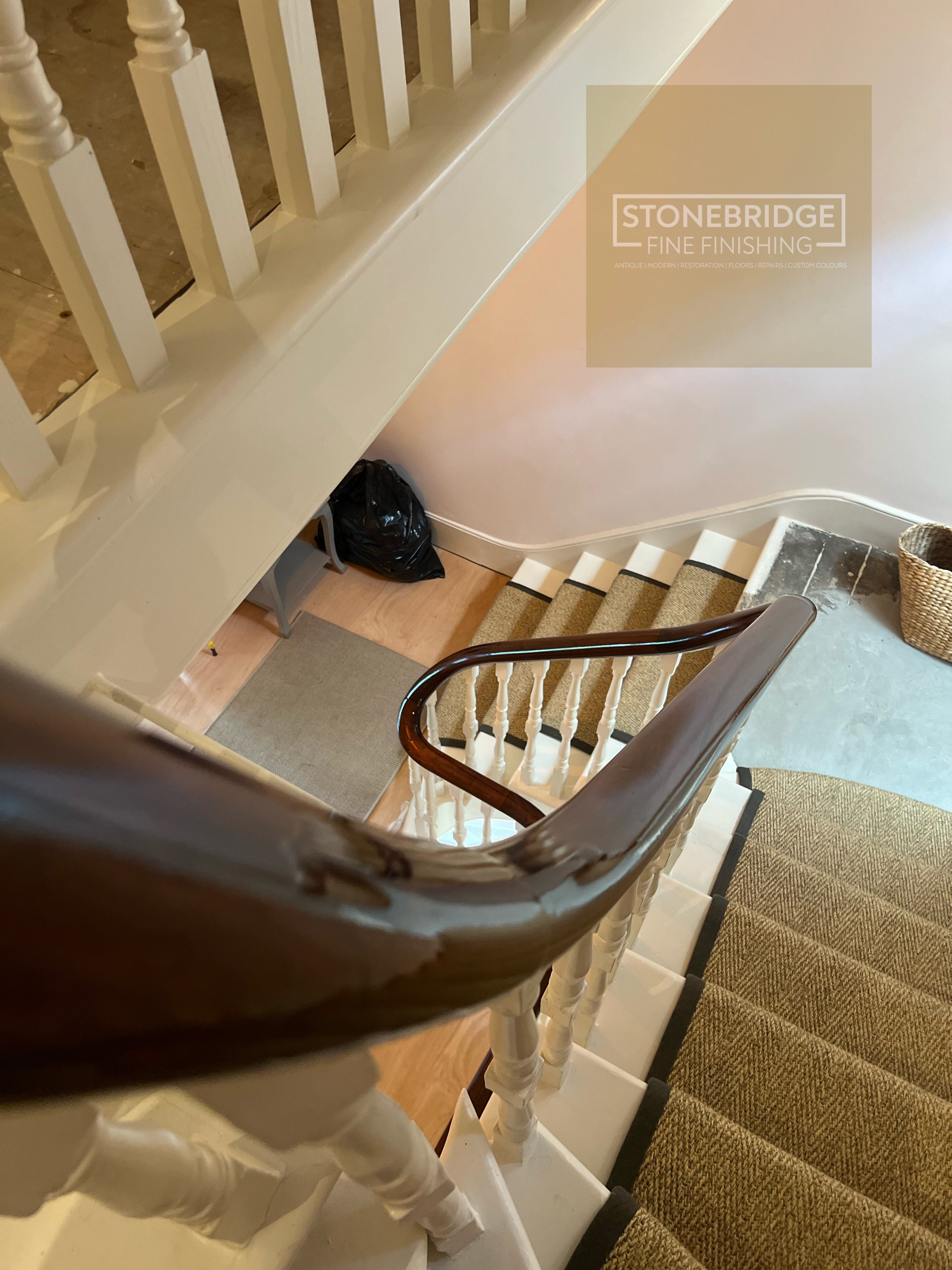 View of a staircase from the top looking down, with a wooden handrail, white balusters, beige carpeted steps, and a beige wall with a logo for Stonebridge Fine Finishing on the wall.