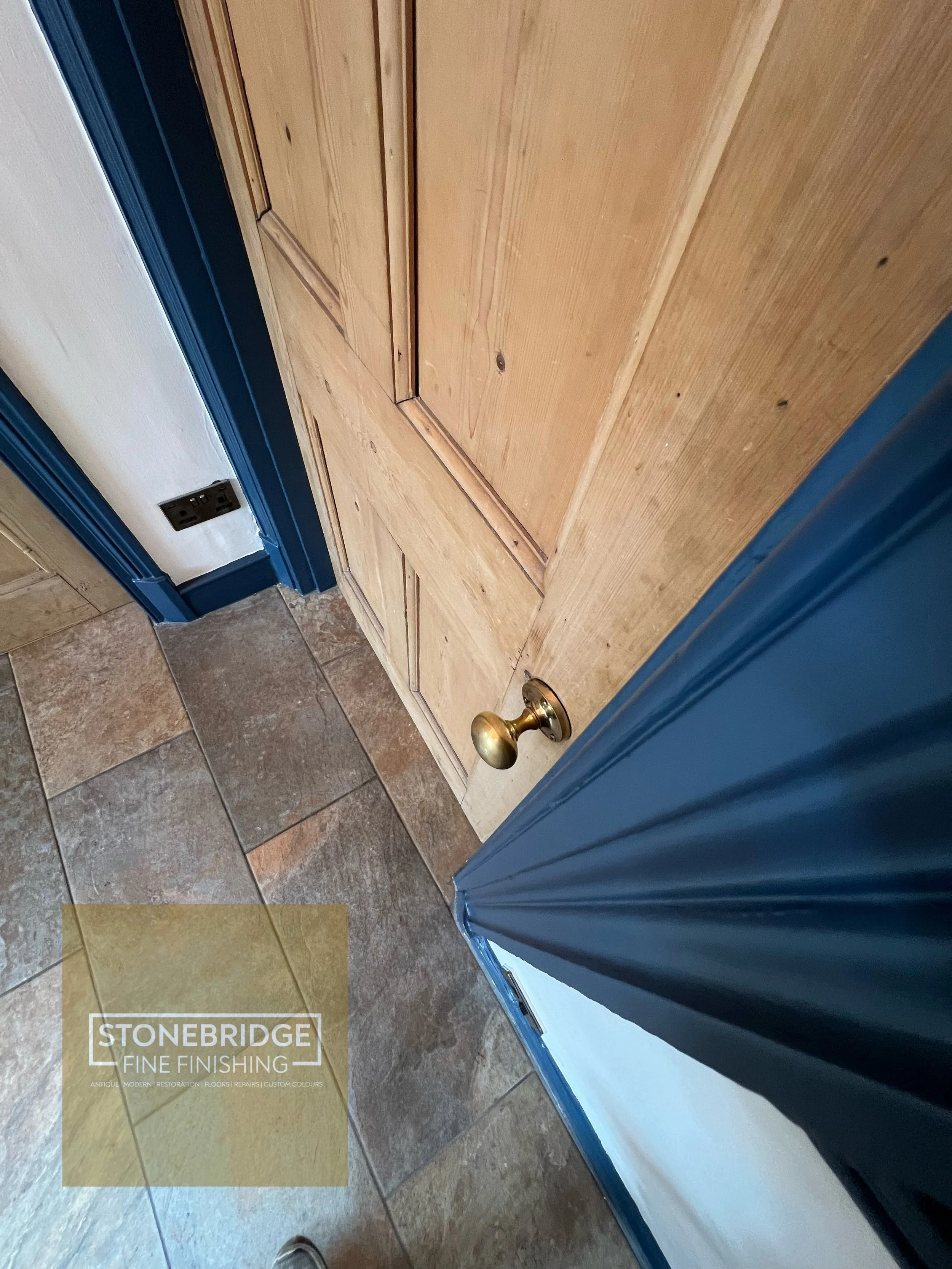 Wooden door with a brass doorknob, blue trim, and a black doorstop on the wall next to it, with tiled flooring and a blue and white wall.