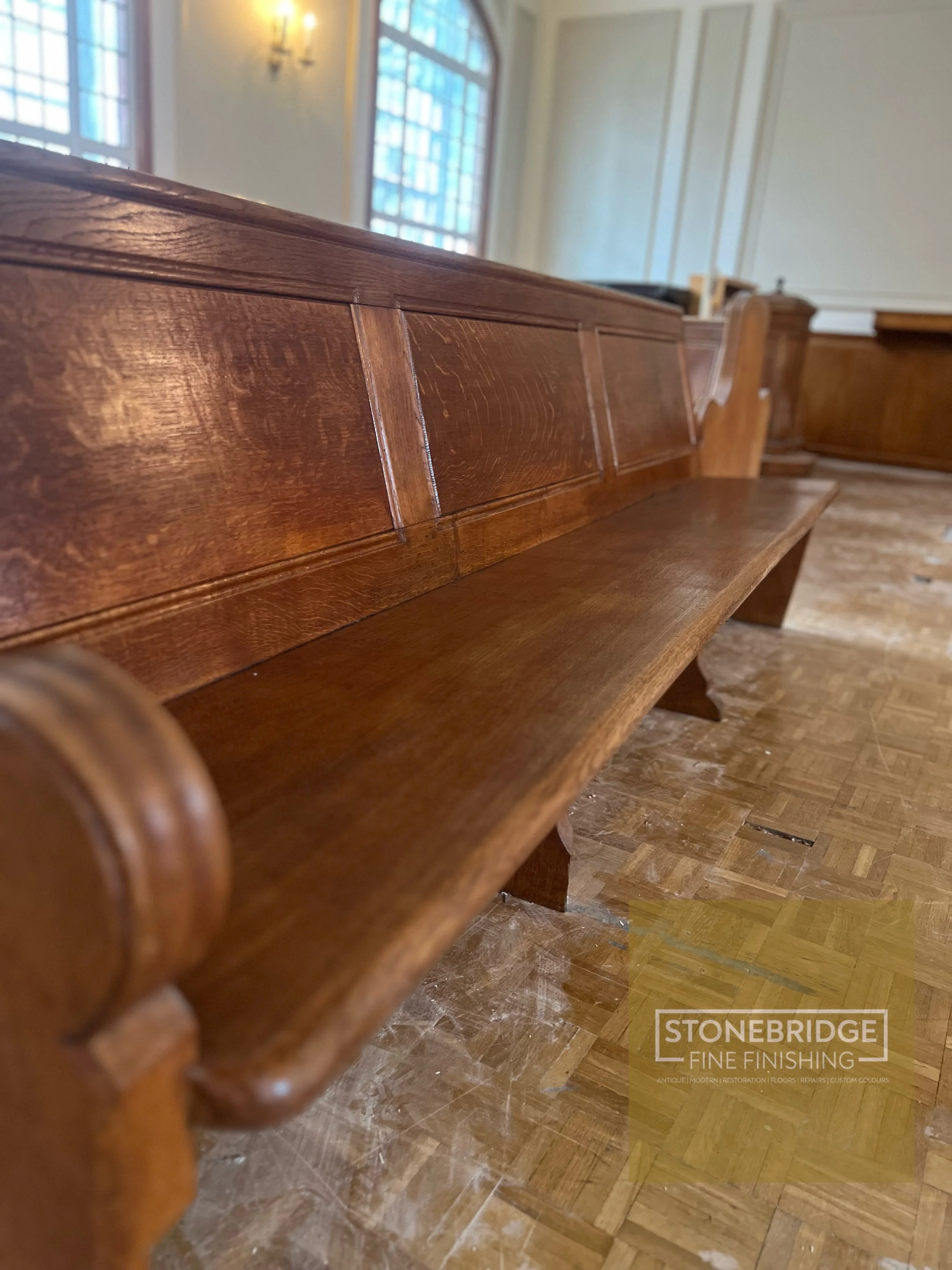 Close-up of a polished wooden pew in a church or similar setting, with a decorative rounded armrest visible in the foreground. Large windows with multiple panes and a wall-mounted light fixture can be seen in the background.