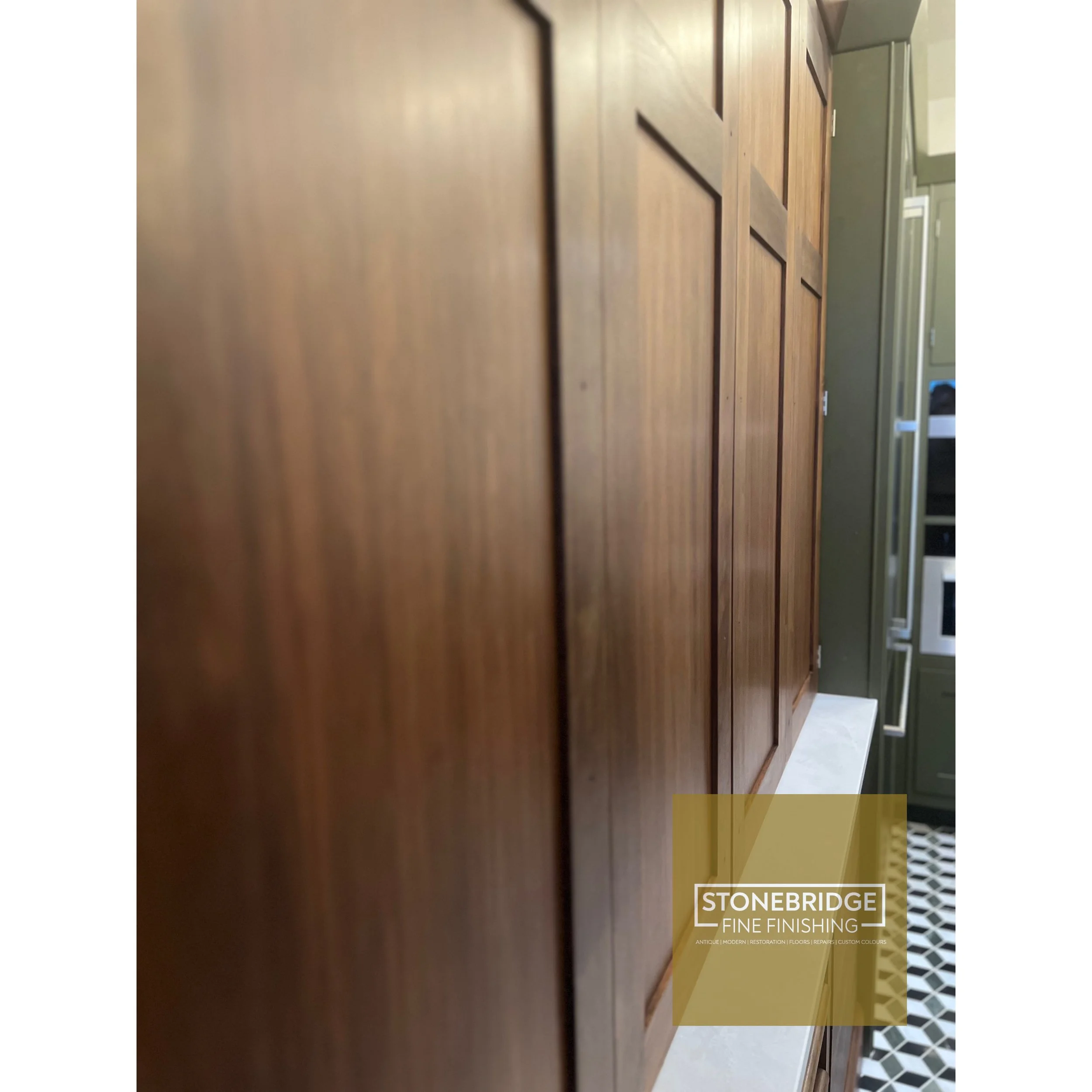 Close-up of a wooden cabinet surface with a small portion of a tile floor visible in the background, and a logo for Stonebridge Fine Finishing at the bottom right corner.