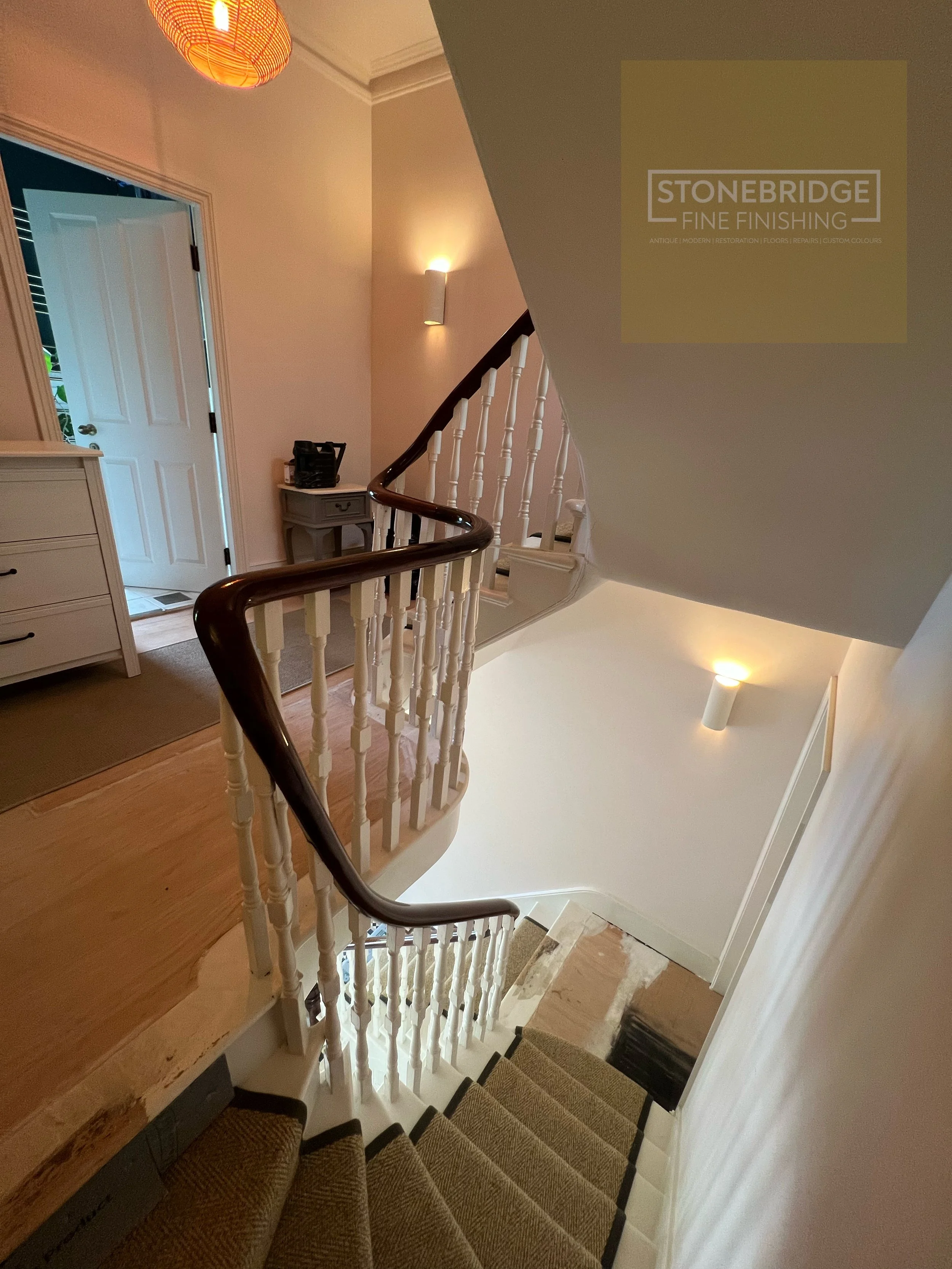 View of a staircase with a curved dark wood handrail, white spindles, carpeted stairs, and a small landing area with a piece of furniture and a basket, leading up to a hallway with a wall sconce and a door.